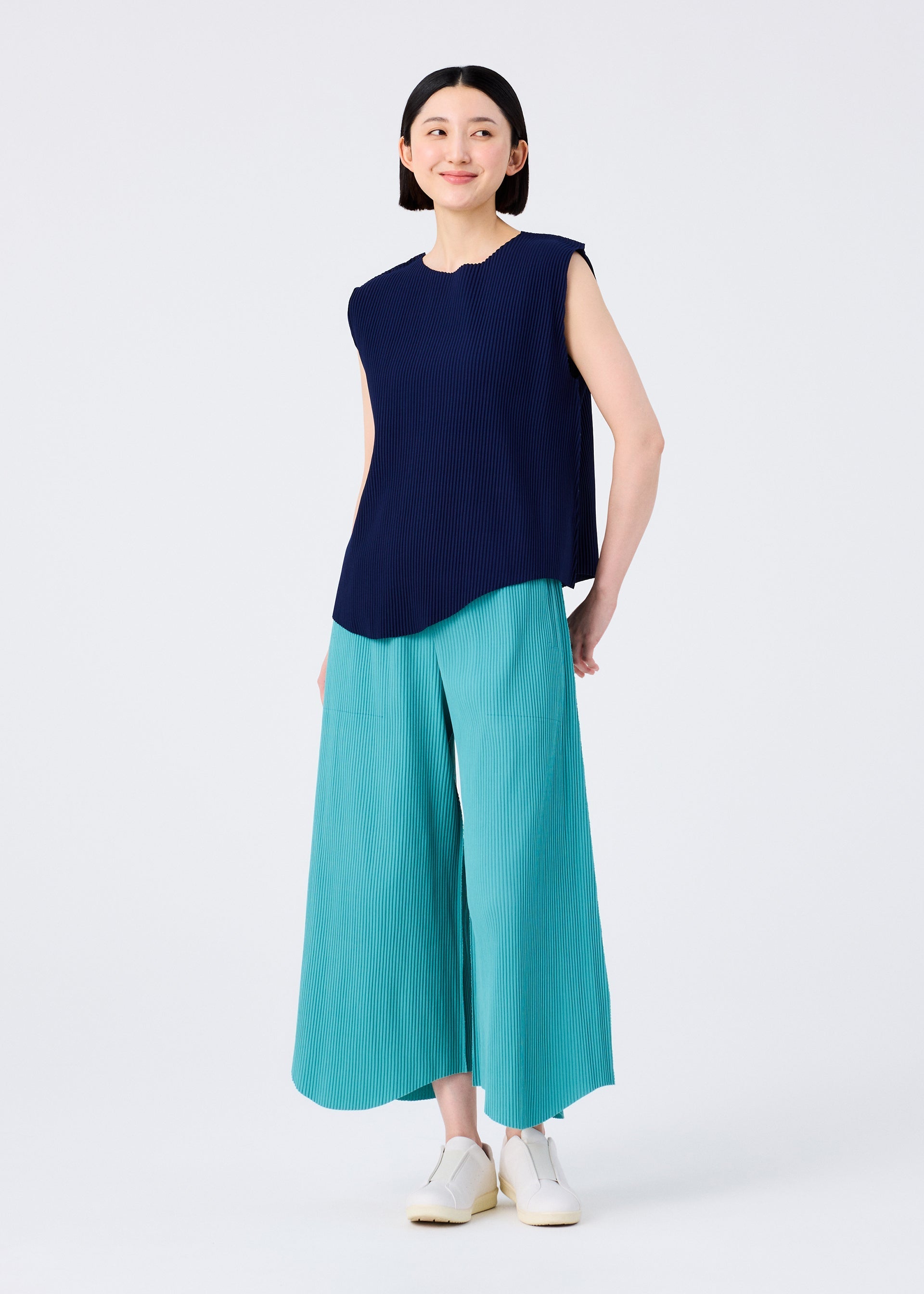Zoomed view A woman with short black hair smiles in a sleeveless navy top and aqua FINE KNIT PLEATS BOTTOM 1 pants by me ISSEY MIYAKE, paired with white shoes against a plain white background.