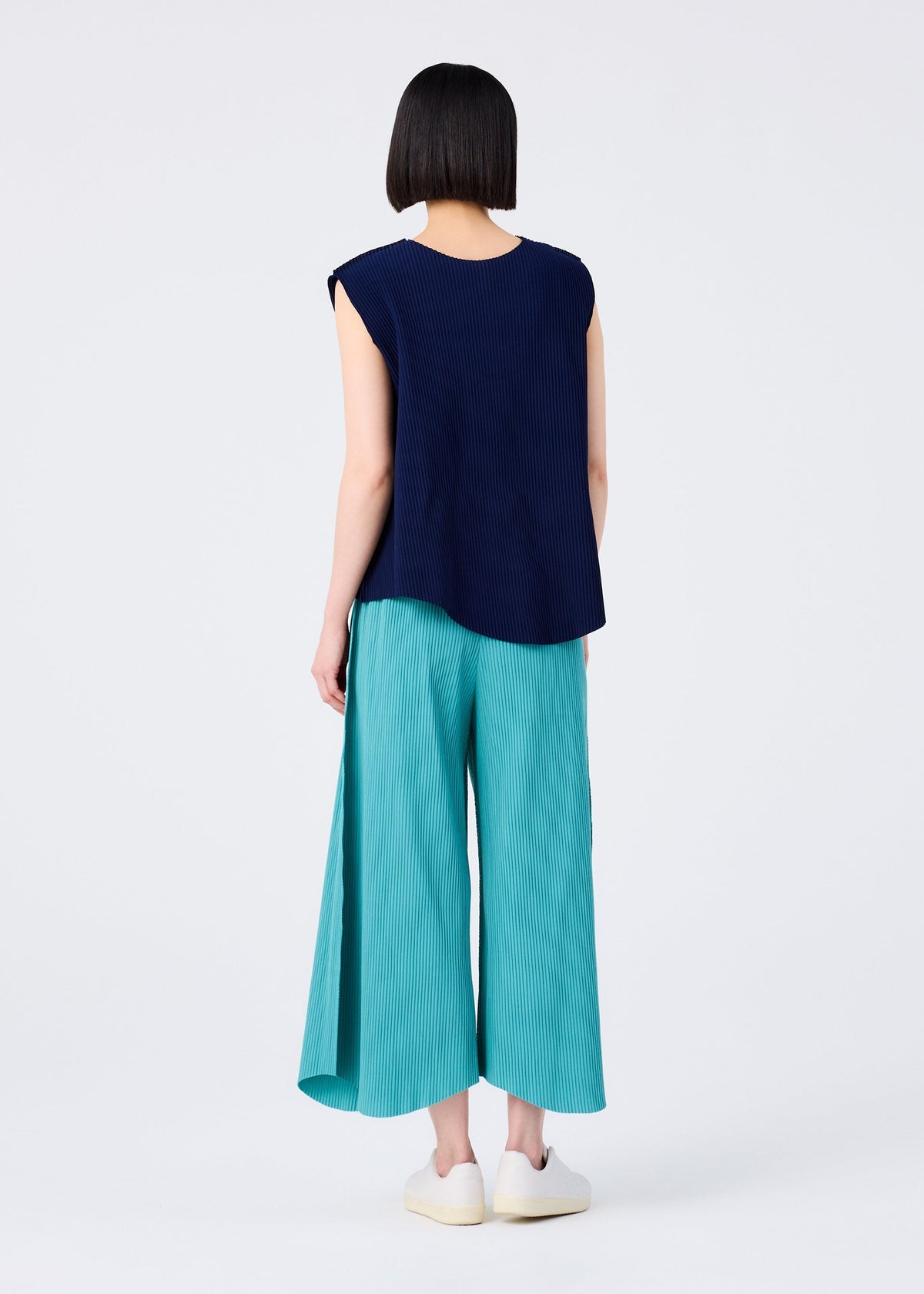 A person with short dark hair stands with their back to the camera, wearing me ISSEY MIYAKE FINE KNIT PLEATS BOTTOM 1, a sleeveless navy top, and white slip-on shoes against a plain white background. view 6 of 6