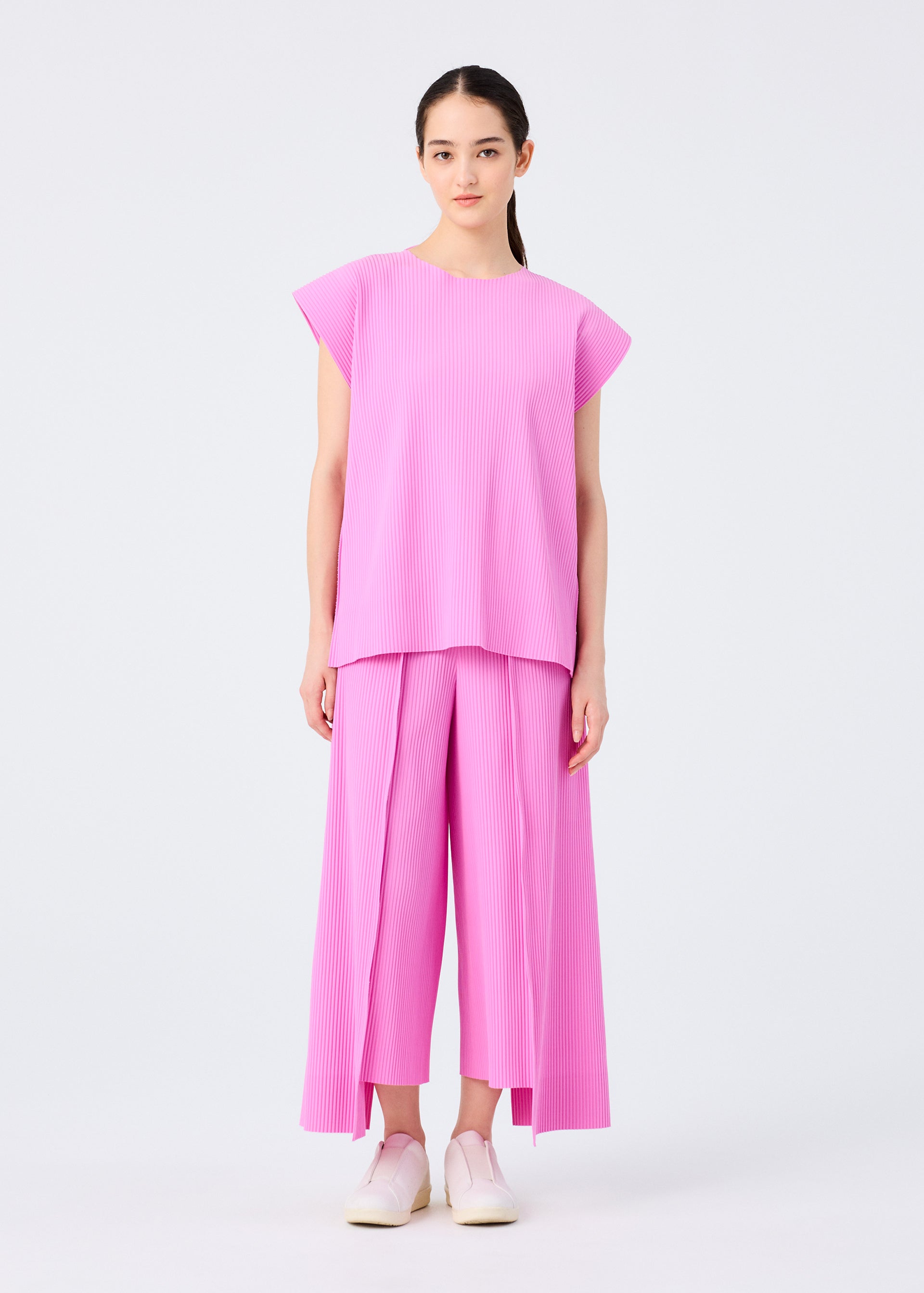 Zoomed view A woman stands facing forward, dressed in the TRICK PLEATS set—loose-fitting, short-sleeved light pink pleated top and wide-leg pants by me ISSEY MIYAKE—paired with white slip-on shoes against a plain white background.