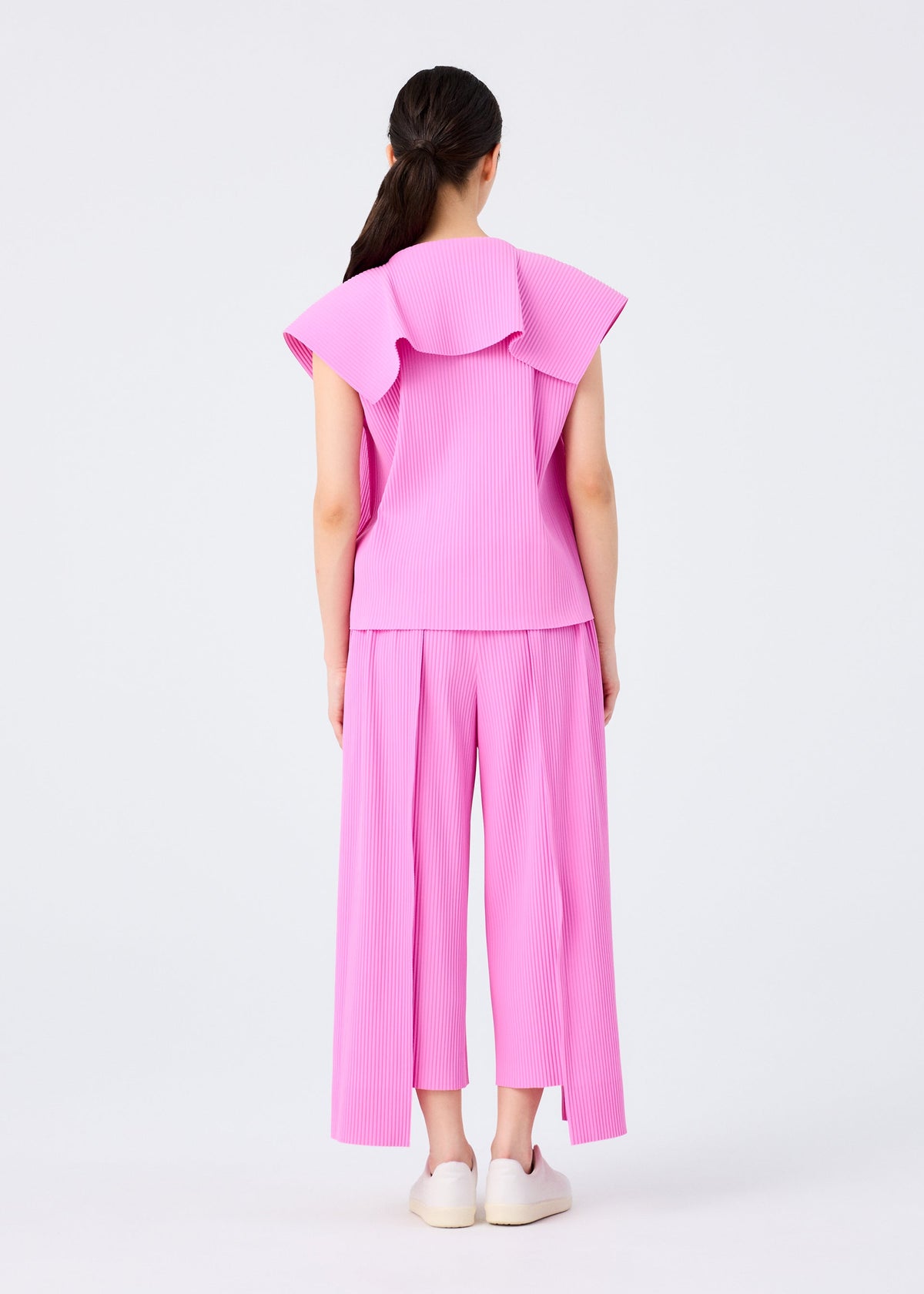 A woman with long dark hair in a ponytail faces away, wearing the me ISSEY MIYAKE TRICK PLEATS set—loose pink sleeveless top with large collar and matching wide-leg fine knit pleated pants—paired with white slip-ons, posed against a plain light background. view 7 of 7