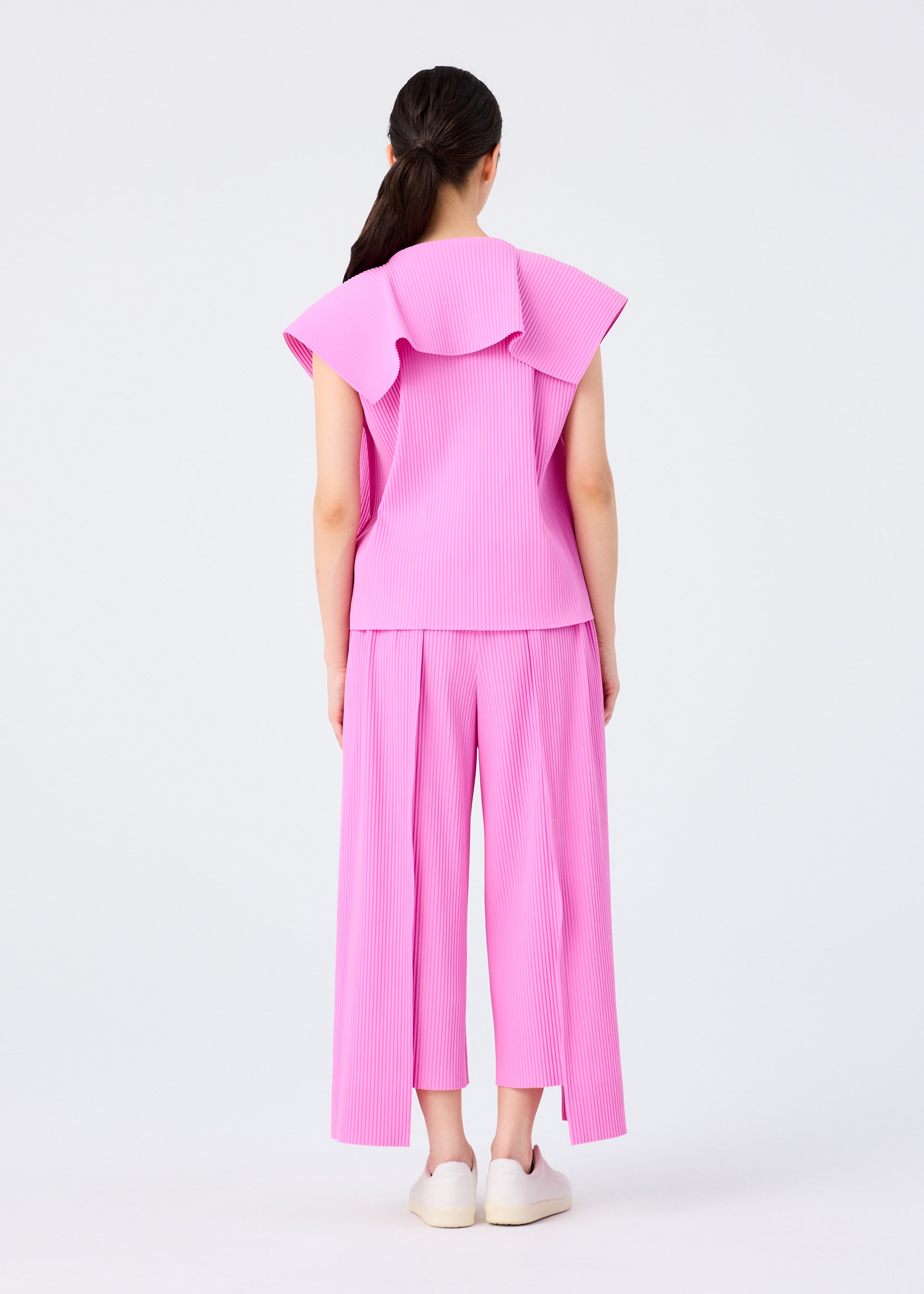 Zoomed view A woman with long dark hair in a ponytail faces away, wearing the me ISSEY MIYAKE TRICK PLEATS set—loose pink sleeveless top with large collar and matching wide-leg fine knit pleated pants—paired with white slip-ons, posed against a plain light background.