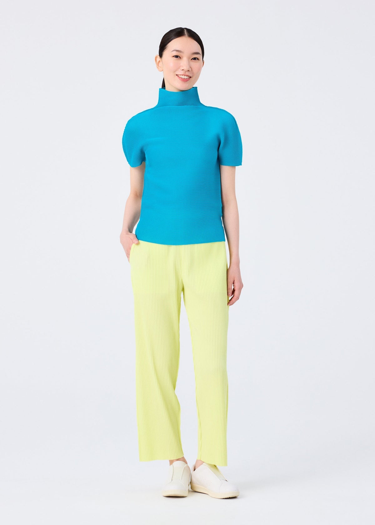 A woman smiles against a white background, wearing the FINE KNIT PLEATS COLOR 2 top and pants by me ISSEY MIYAKE in bright blue and light yellow, paired with white shoes and one hand in her pocket. view 6 of 7