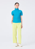 A woman smiles against a white background, wearing the FINE KNIT PLEATS COLOR 2 top and pants by me ISSEY MIYAKE in bright blue and light yellow, paired with white shoes and one hand in her pocket.