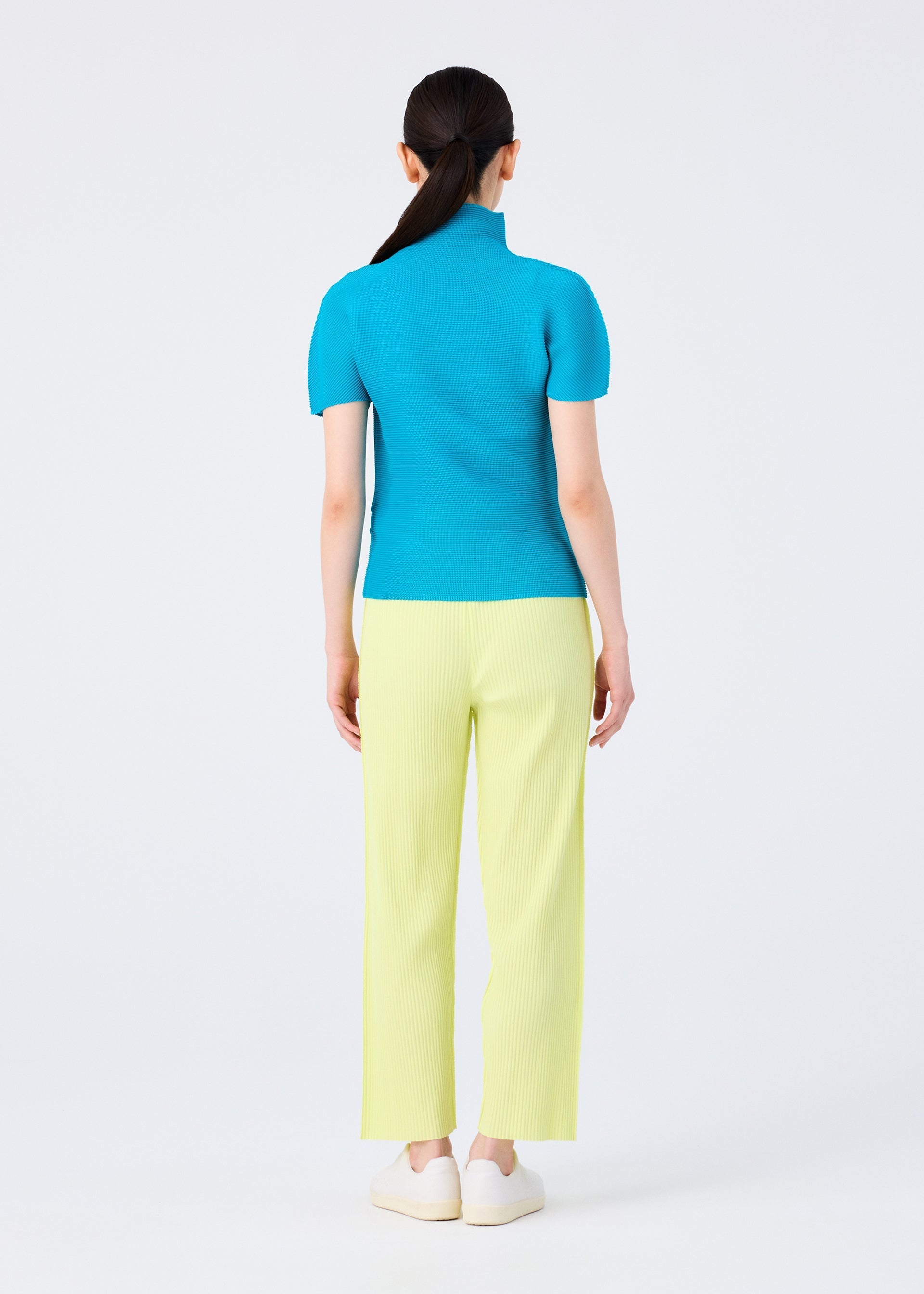 Zoomed view A woman with straight dark hair pulled back stands against a white background, wearing me ISSEY MIYAKE's FINE KNIT PLEATS COLOR 2 wide-leg pants in pale yellow, a bright blue lightweight short-sleeve top, and white shoes.