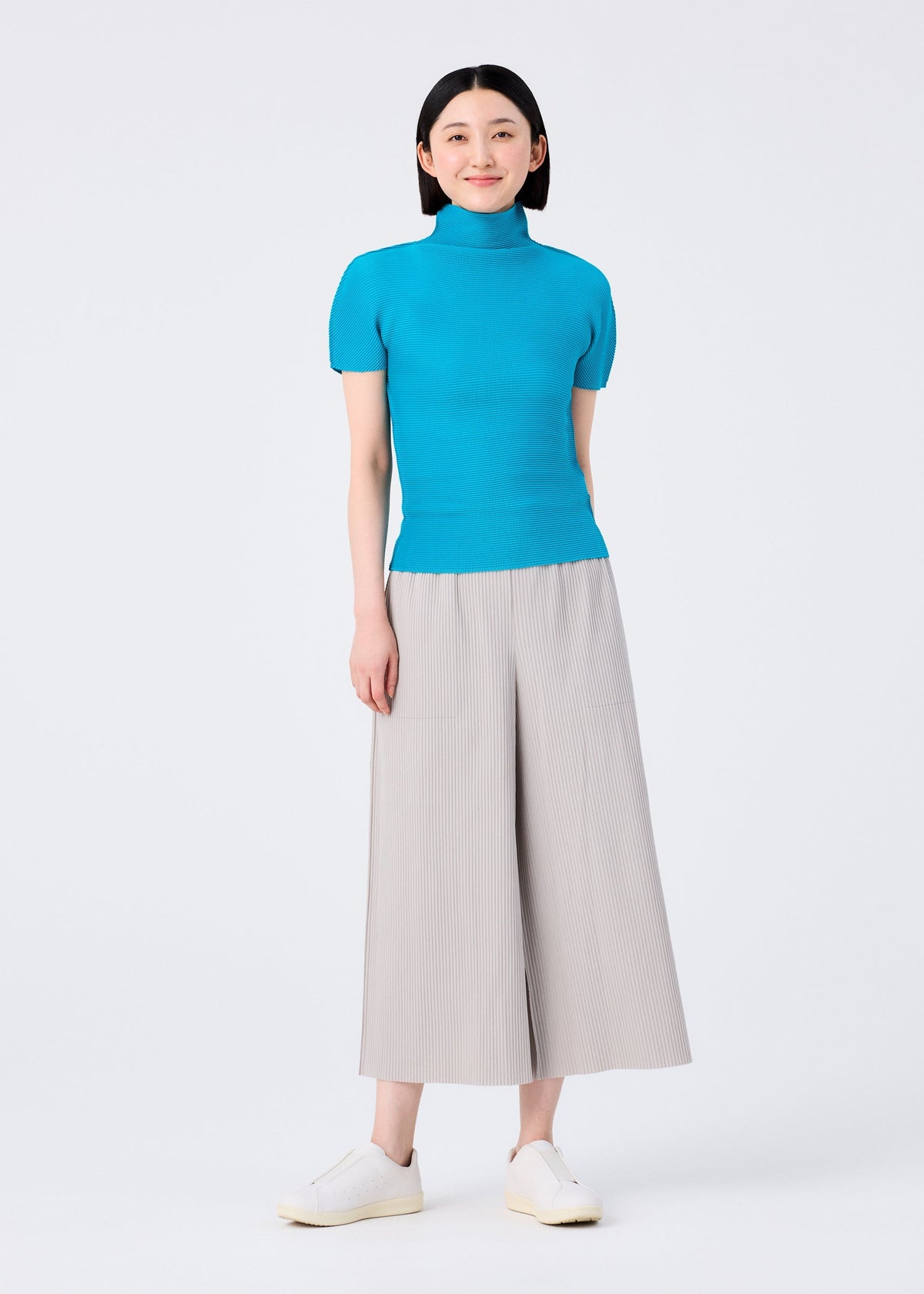 A woman smiles in the me ISSEY MIYAKE FINE KNIT PLEATS COLOR 2 beige pleated wide-leg cropped pants, a bright blue short-sleeve turtleneck, and white slip-on shoes, standing against a plain white background. view 6 of 7