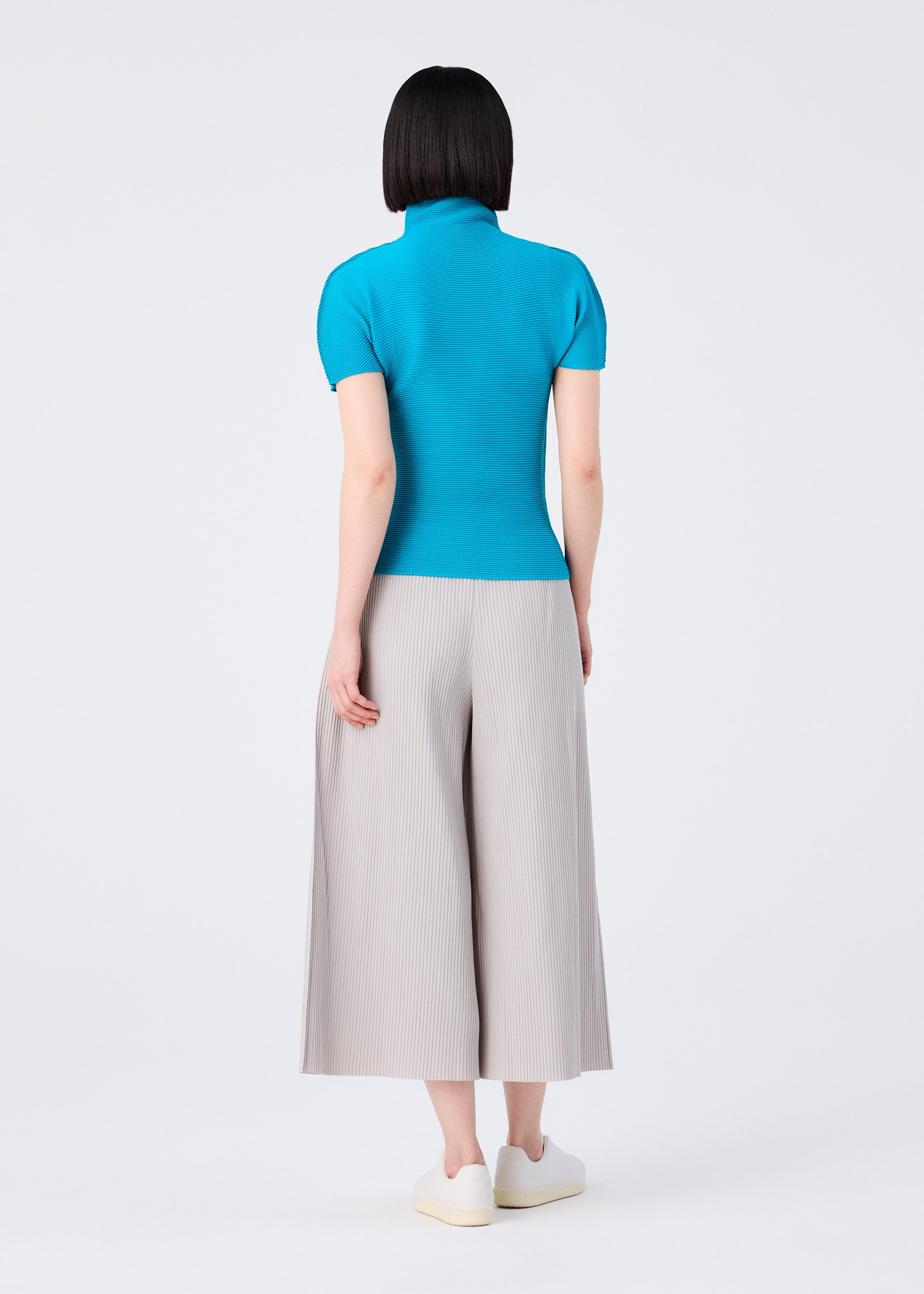 Zoomed view A person with straight, chin-length black hair stands facing away, wearing the me ISSEY MIYAKE FINE KNIT PLEATS COLOR 2 light gray pants, a bright blue short-sleeve turtleneck top, and white slip-on shoes against a plain white background.