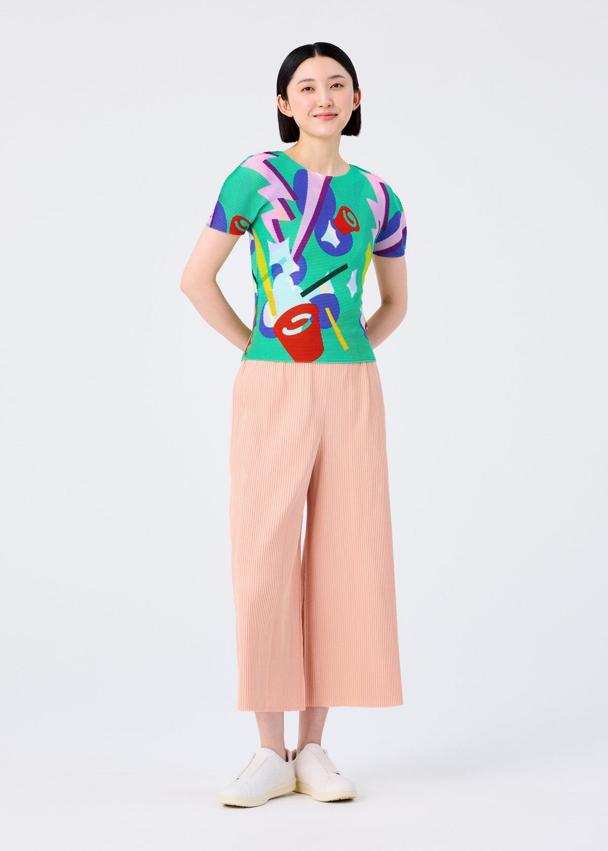 A person faces forward, wearing a colorful abstract-patterned short-sleeve top, me ISSEY MIYAKE FINE KNIT PLEATS BOTTOM 2 wide-legged peach pants, and white shoes, against a plain light background. view 6 of 8