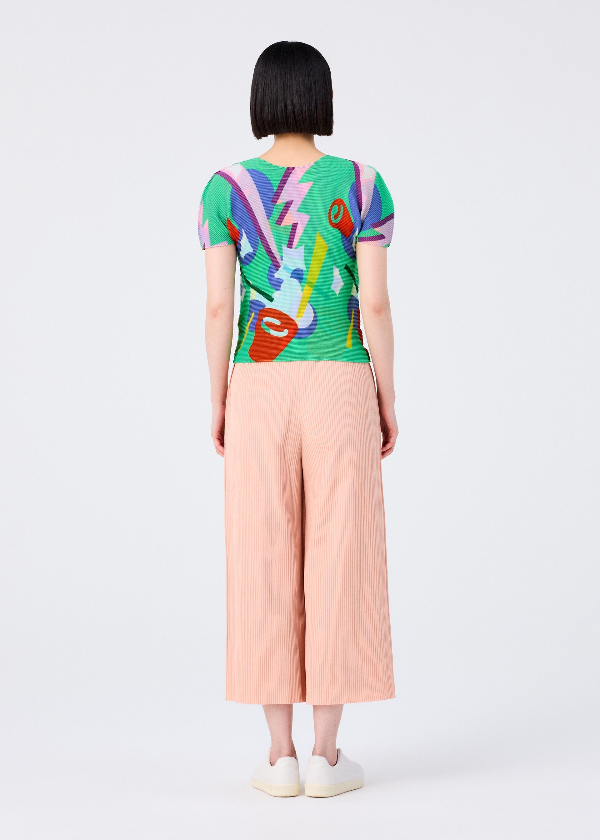Zoomed view A person with short black hair, seen from behind, wears a vibrant top and light pink FINE KNIT PLEATS BOTTOM 2 pants by me ISSEY MIYAKE, plus white slip-on shoes, standing against a plain white background.