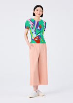A woman stands smiling with hands in pockets, wearing a colorful abstract short-sleeve top, me ISSEY MIYAKE FINE KNIT PLEATS BOTTOM 2 pants in light peach, and white shoes against a plain white background.