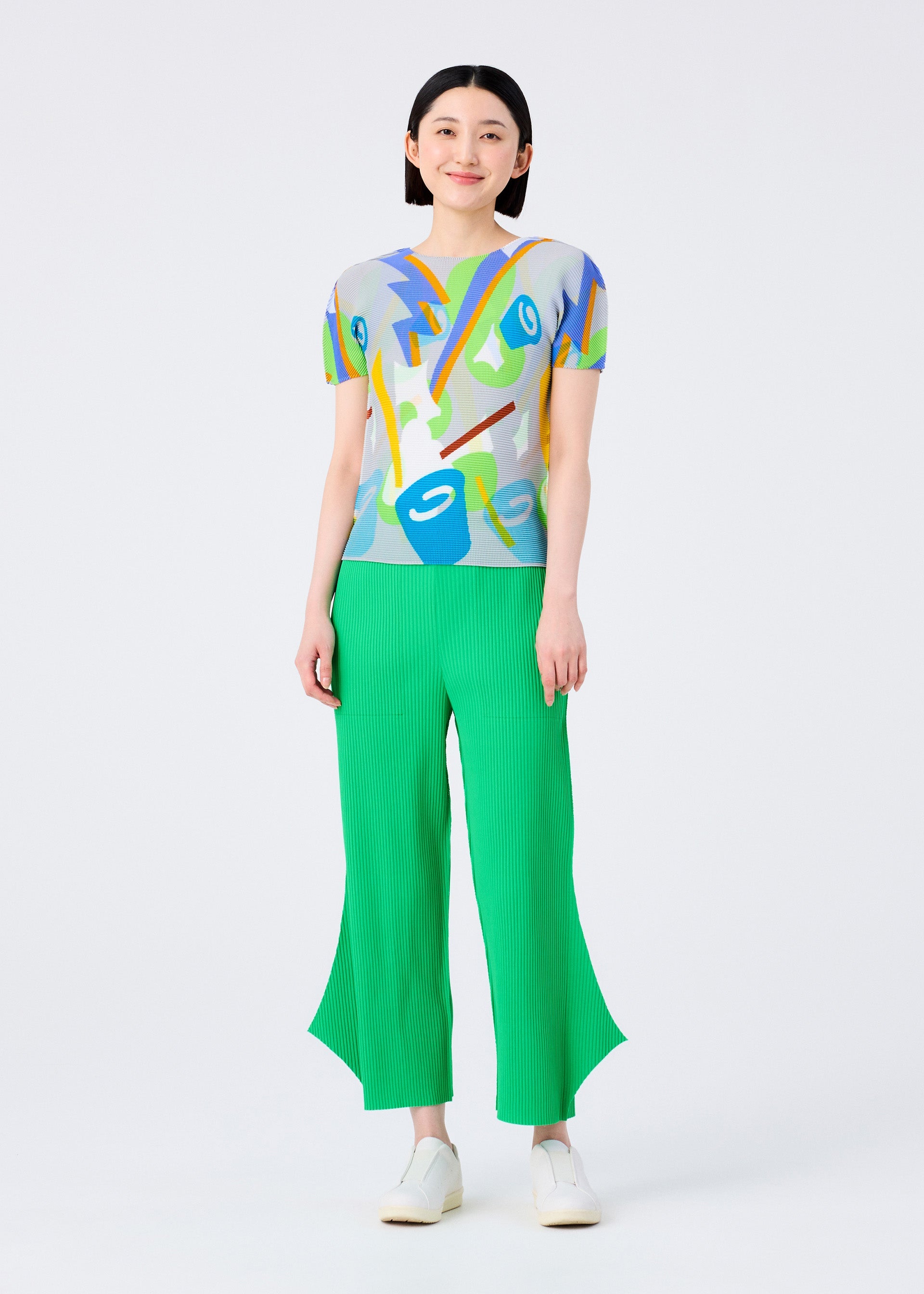 Zoomed view A woman stands smiling against a plain white background, wearing bright green FINE KNIT PLEATS BOTTOM 2 pants by me ISSEY MIYAKE, a colorful abstract print top, and white shoes.