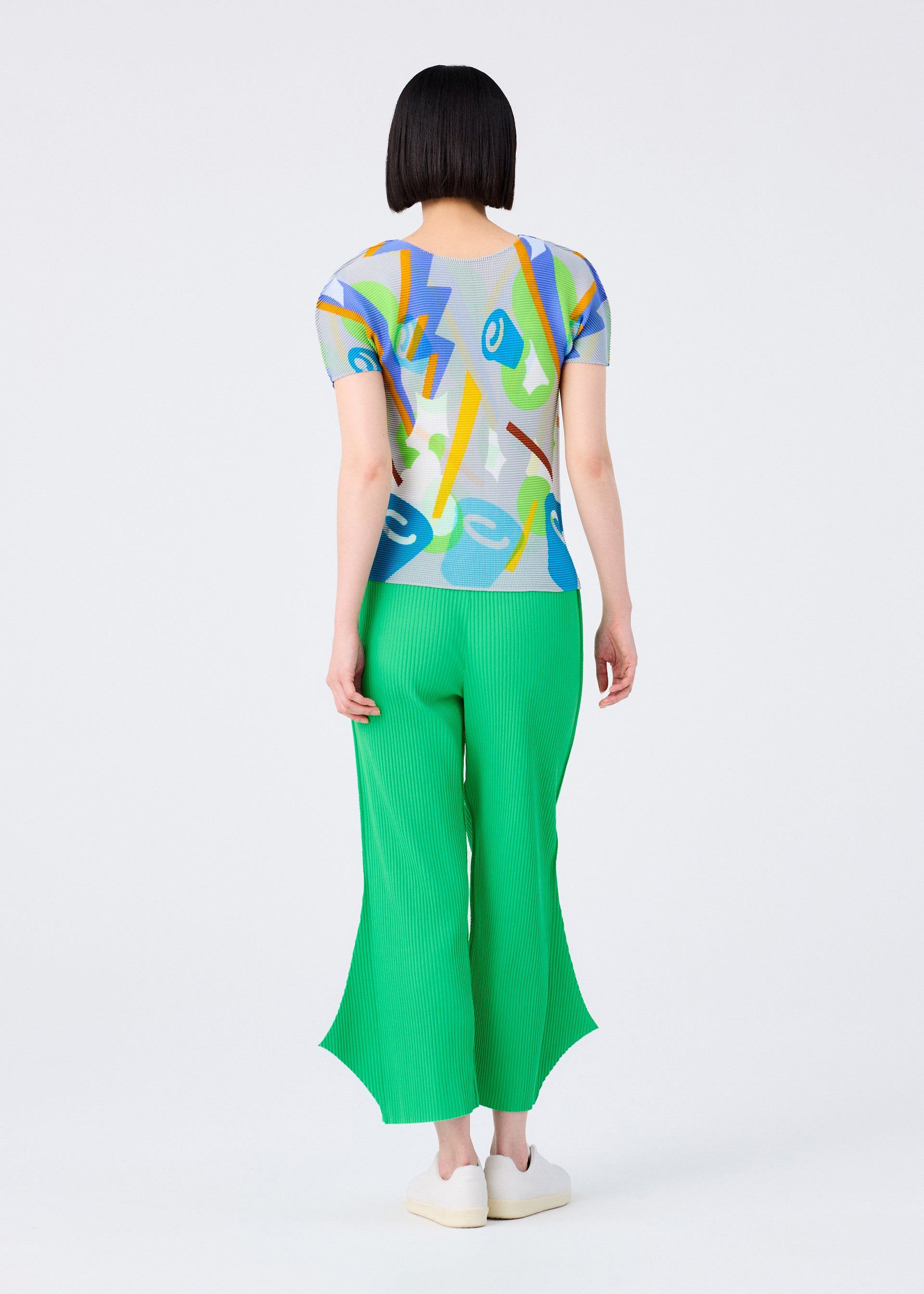 Zoomed view A person with short black hair stands facing away, wearing a colorful abstract top and bright green FINE KNIT PLEATS BOTTOM 2 pants by me ISSEY MIYAKE, paired with white shoes, on a plain light background.