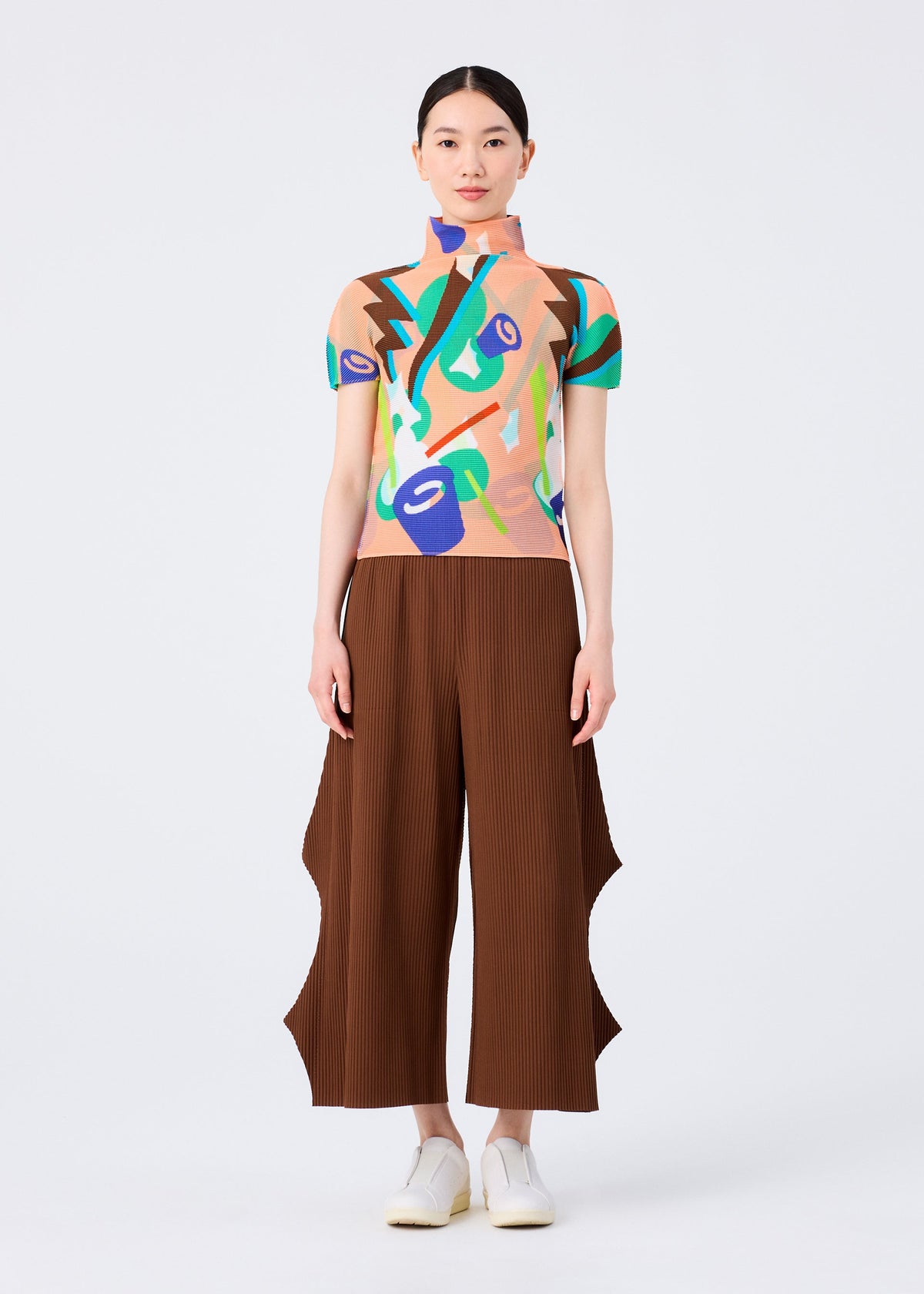 A woman faces forward against a white background, wearing a colorful abstract-patterned short-sleeve top, me ISSEY MIYAKE FINE KNIT PLEATS BOTTOM 2 wide-leg pants with wavy side slits, and white shoes. view 5 of 6