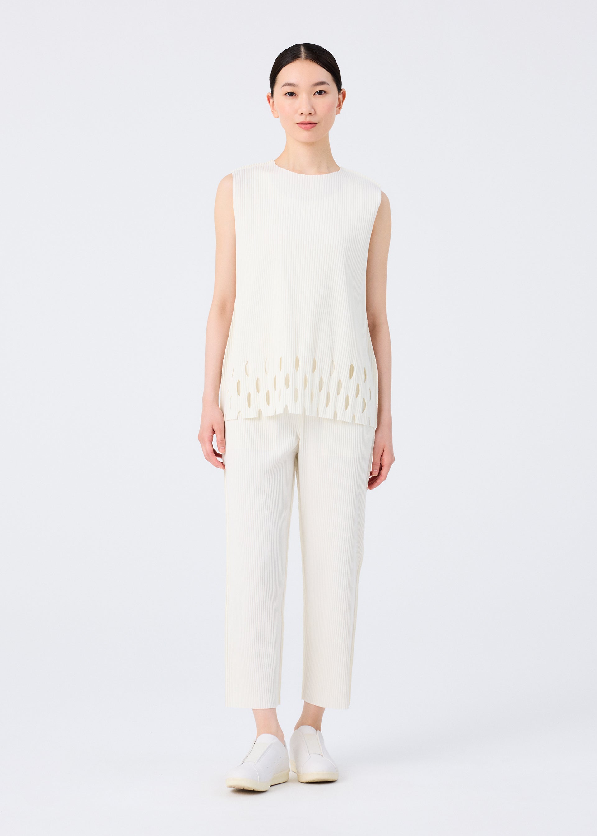 Zoomed view A woman with straight dark hair pulled back wears me ISSEY MIYAKE's FINE KNIT PLEATS BOTTOM 3 pants, a white sleeveless ribbed top with oval cutouts at the hem, and white slip-on shoes against a plain white background.