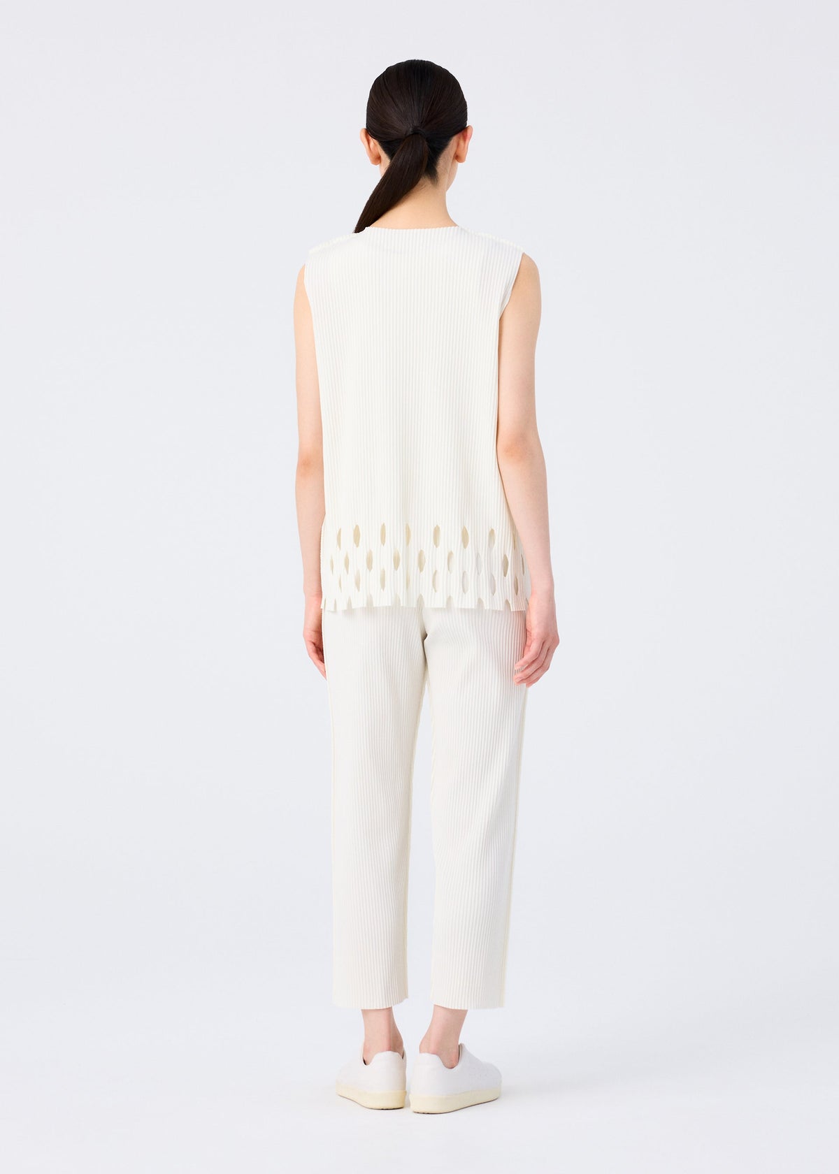 A person with long dark hair in a low ponytail faces away, wearing me ISSEY MIYAKE's FINE KNIT PLEATS BOTTOM 3—straight white pants styled with a sleeveless white top and slip-on shoes against a plain white background. view 5 of 5