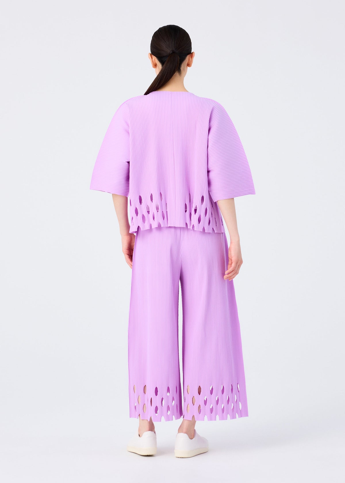 A person with long dark hair in a low ponytail wears the me ISSEY MIYAKE FINE KNIT PLEATS BOTTOM 3 pants, featuring oval cutouts at the hems, paired with a loose pleated top and white shoes against a plain white background. view 6 of 7
