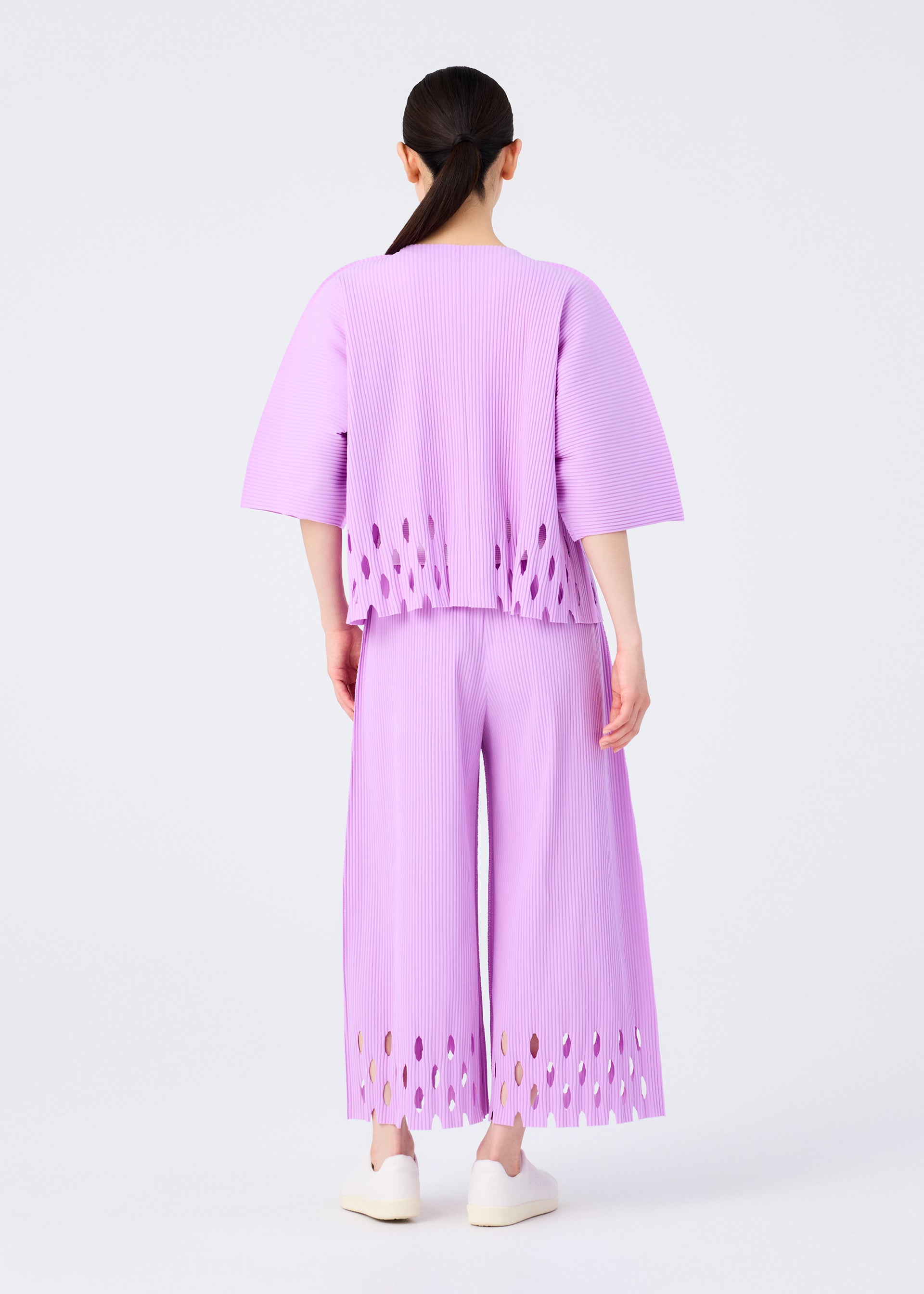 Zoomed view A person with long dark hair in a low ponytail wears the me ISSEY MIYAKE FINE KNIT PLEATS BOTTOM 3 pants, featuring oval cutouts at the hems, paired with a loose pleated top and white shoes against a plain white background.