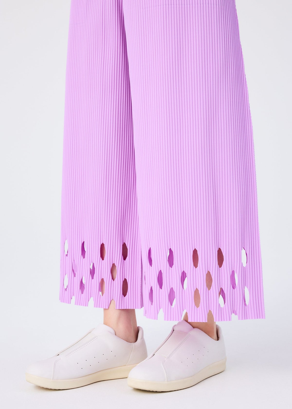A person wears me ISSEY MIYAKE's FINE KNIT PLEATS BOTTOM 3 pants with a scalloped cut-out hem, paired with pale pink slip-on sneakers, standing against a plain light background. view 7 of 7