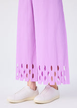 A person wears me ISSEY MIYAKE's FINE KNIT PLEATS BOTTOM 3 pants with a scalloped cut-out hem, paired with pale pink slip-on sneakers, standing against a plain light background.