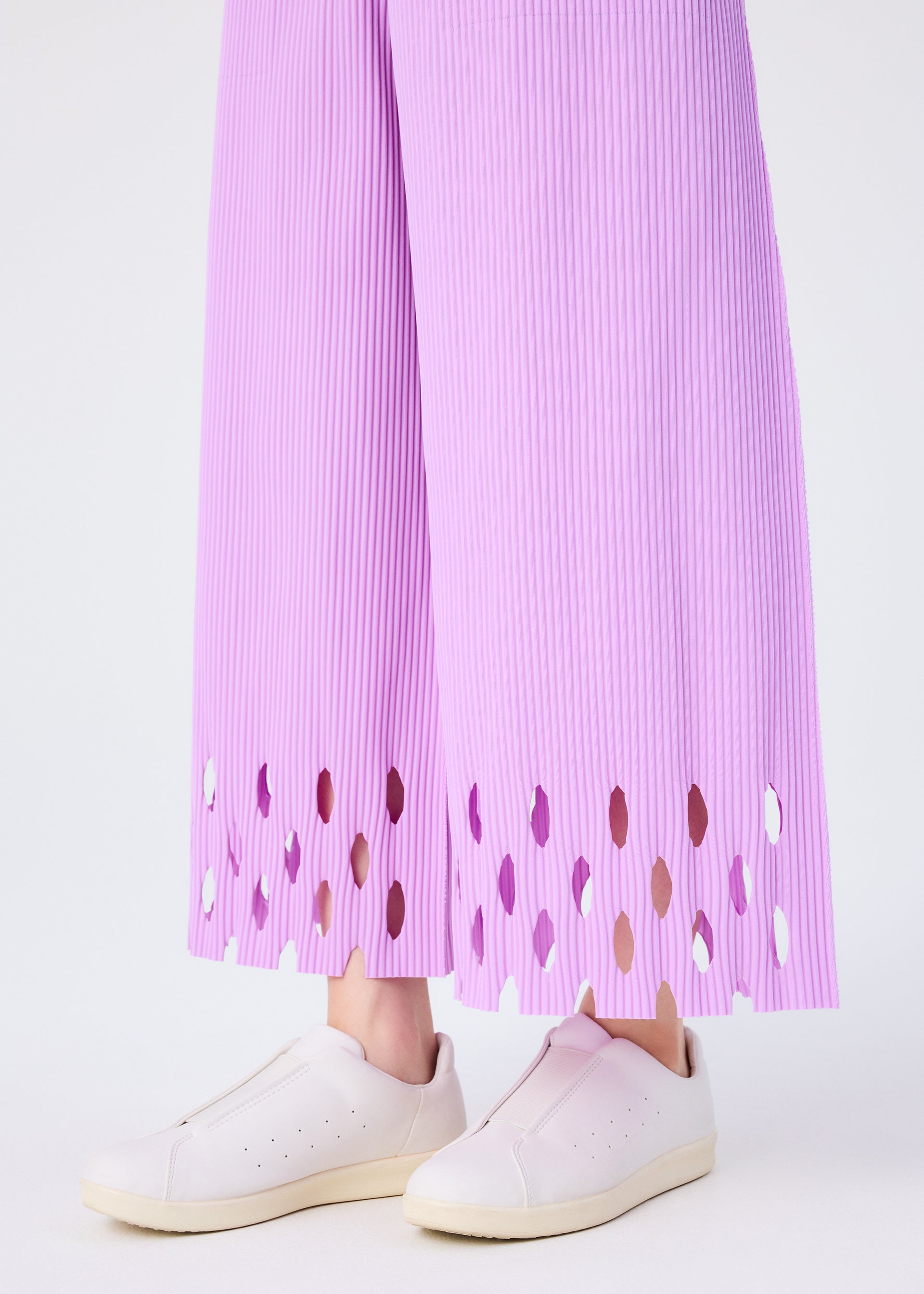 Zoomed view A person wears me ISSEY MIYAKE's FINE KNIT PLEATS BOTTOM 3 pants with a scalloped cut-out hem, paired with pale pink slip-on sneakers, standing against a plain light background.