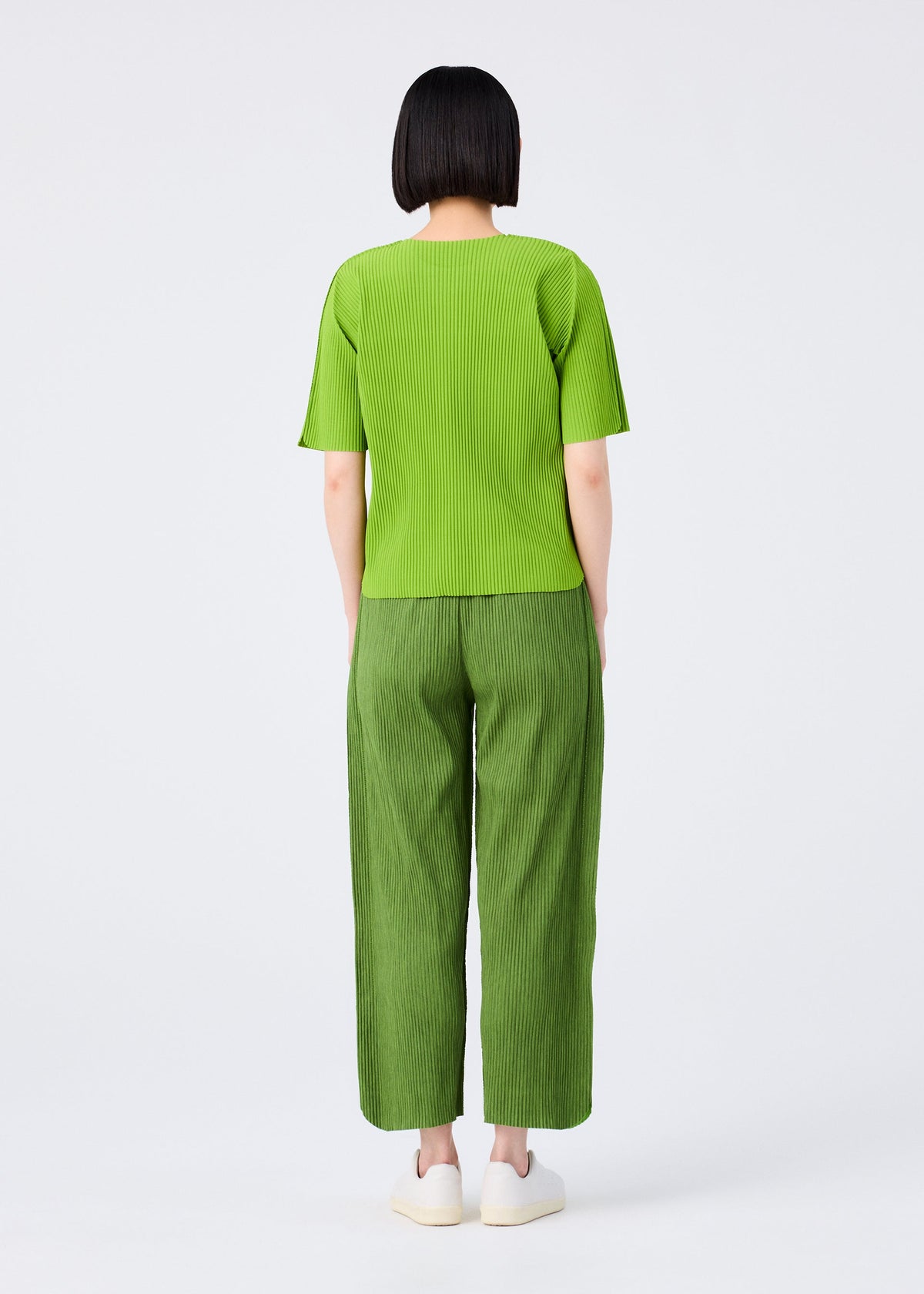 A person with short black hair stands facing away, wearing a bright green top, me ISSEY MIYAKE MIX FINE KNIT PLEATS BOTTOM pants in matching green, and white shoes against a plain white background. view 7 of 7