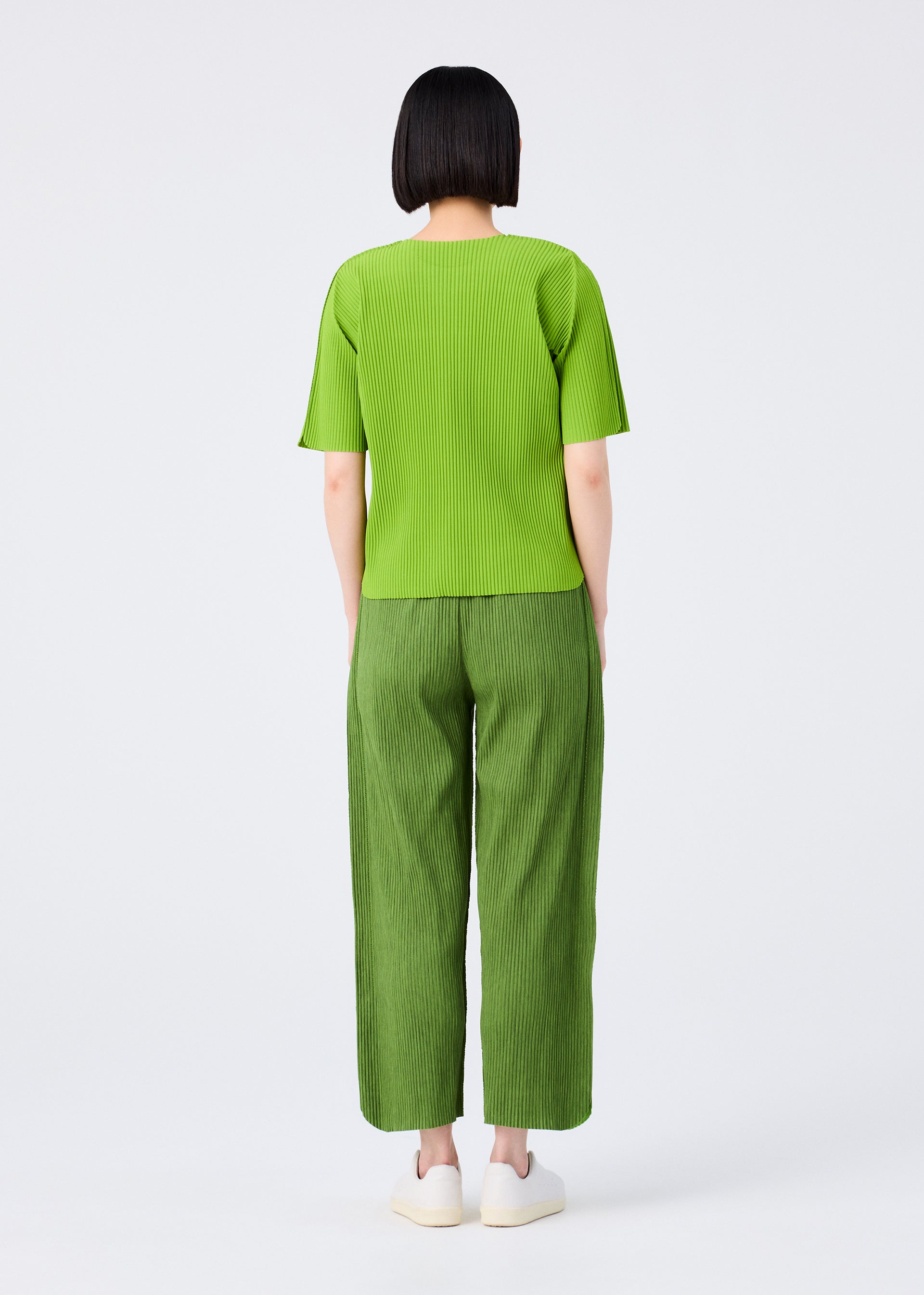 Zoomed view A person with short black hair stands facing away, wearing a bright green top, me ISSEY MIYAKE MIX FINE KNIT PLEATS BOTTOM pants in matching green, and white shoes against a plain white background.