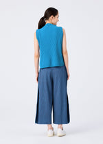 A woman faces away, dark hair in a low ponytail, wearing me ISSEY MIYAKE’s MIX FINE KNIT PLEATS BOTTOM—lightweight wide pants with slits and fine knit pleats—paired with a blue sleeveless top and white slip-on shoes against a plain background.