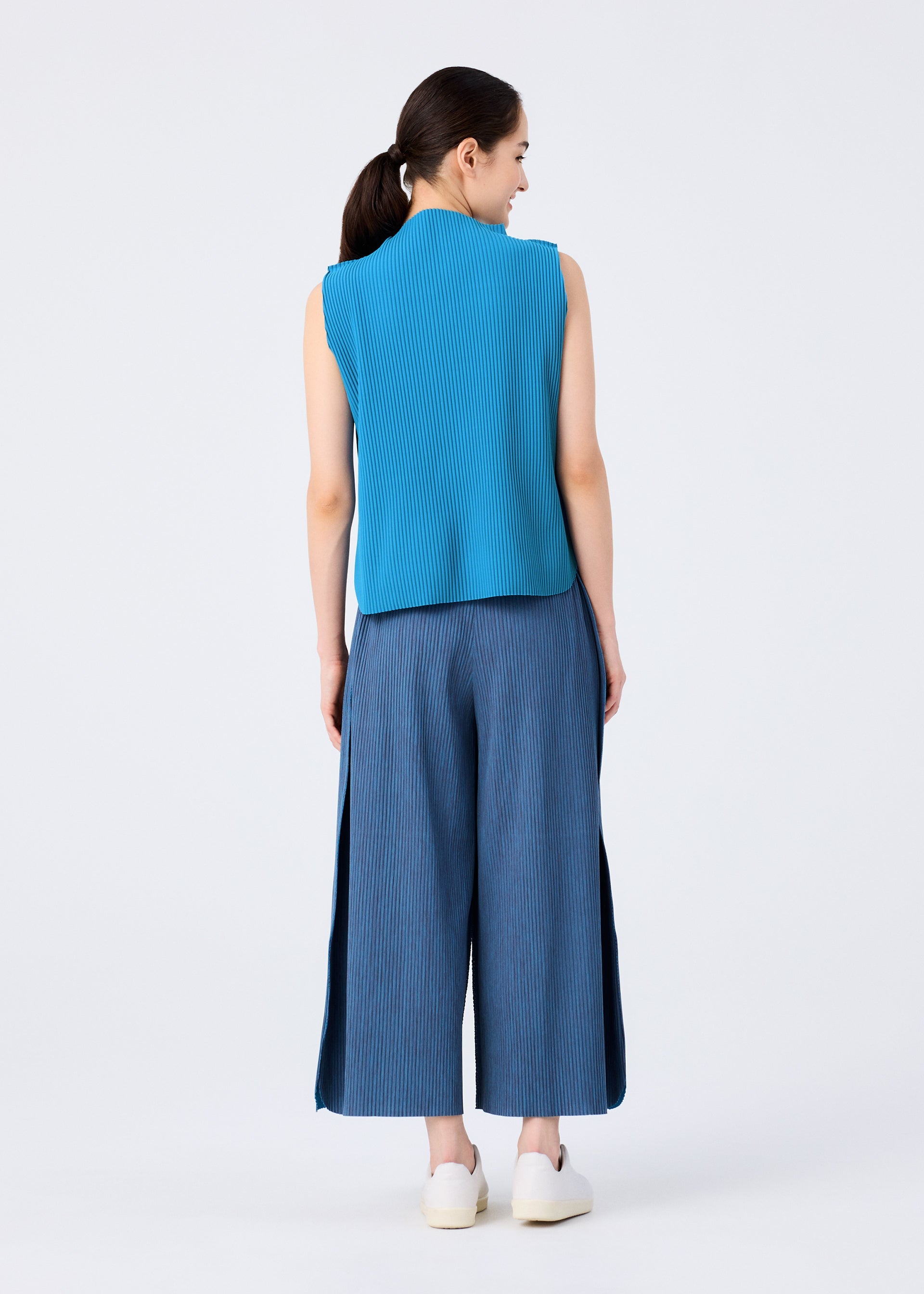 Zoomed view A woman faces away, dark hair in a low ponytail, wearing me ISSEY MIYAKE’s MIX FINE KNIT PLEATS BOTTOM—lightweight wide pants with slits and fine knit pleats—paired with a blue sleeveless top and white slip-on shoes against a plain background.