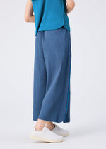 Only the lower half of a person is visible, standing on a white background and wearing me ISSEY MIYAKE MIX FINE KNIT PLEATS BOTTOM in blue, styled with a teal pleated top and white sneakers.