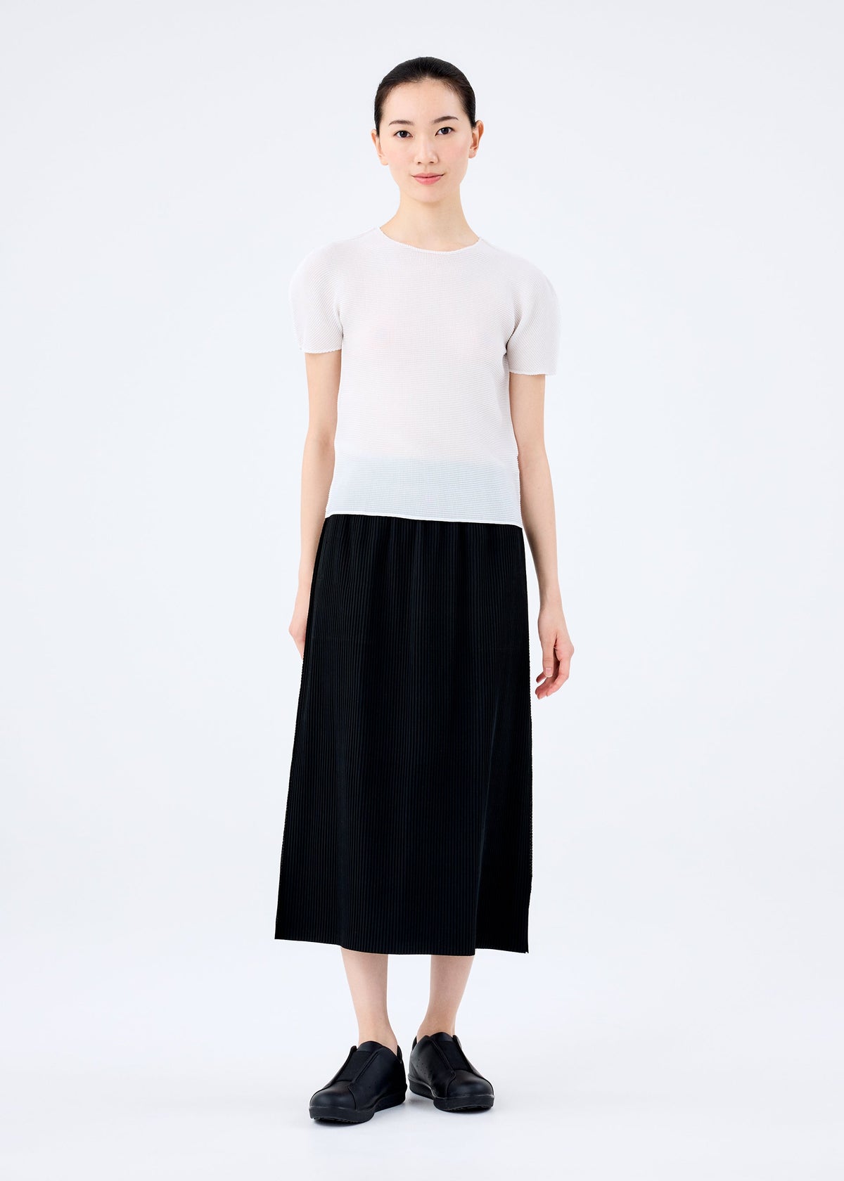 A woman stands against a white background, wearing the me ISSEY MIYAKE FINE KNIT PLEATS BLACK skirt paired with a short-sleeve white top and matching shoes. She faces forward with a neutral expression, her hair pulled back. view 4 of 6