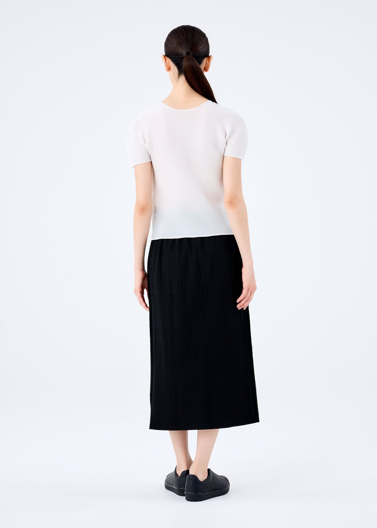 A woman with long dark hair tied back stands facing away from the camera, wearing a me ISSEY MIYAKE FINE KNIT PLEATS BLACK skirt, a short-sleeve white top, and black shoes against a plain light background. view 5 of 6