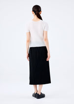 A woman with long dark hair tied back stands facing away from the camera, wearing a me ISSEY MIYAKE FINE KNIT PLEATS BLACK skirt, a short-sleeve white top, and black shoes against a plain light background.