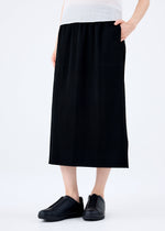 A person stands against a plain white background wearing the FINE KNIT PLEATS BLACK skirt by me ISSEY MIYAKE, paired with a white top and black slip-on shoes, with one hand in their pocket.