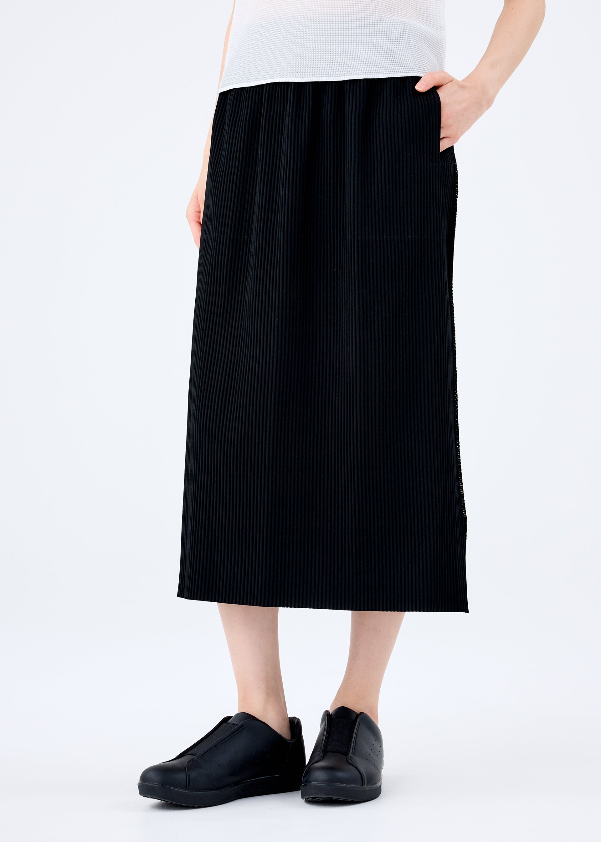 Zoomed view A person stands against a plain white background wearing the FINE KNIT PLEATS BLACK skirt by me ISSEY MIYAKE, paired with a white top and black slip-on shoes, with one hand in their pocket.
