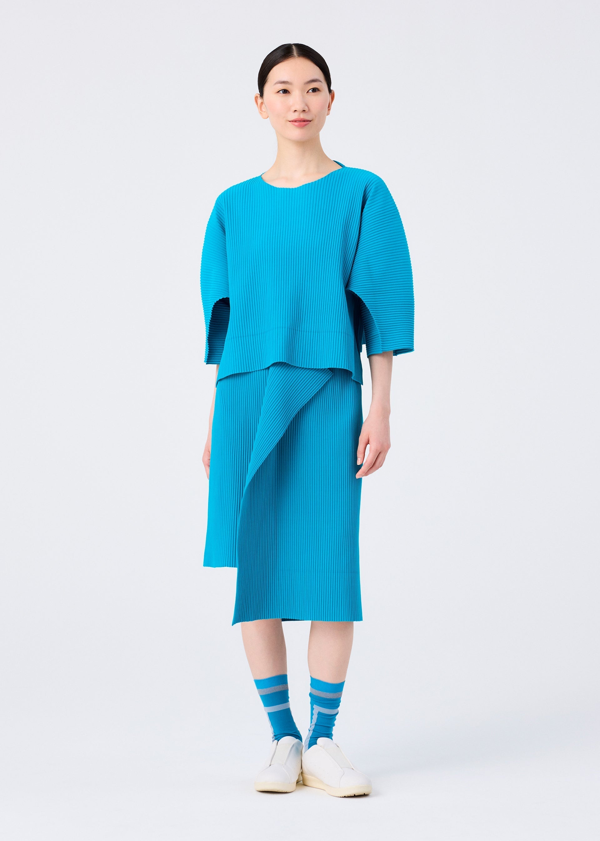 Zoomed view A person wears the TRICK PLEATS skirt and matching top by me ISSEY MIYAKE, featuring a bright blue fine knit with asymmetrical design for a 3D silhouette, paired with blue striped socks and white slip-on shoes.