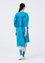 A person with long dark hair stands facing away in the TRICK PLEATS bright blue skirt and matching top by me ISSEY MIYAKE, paired with blue socks and white shoes against a plain white background.
