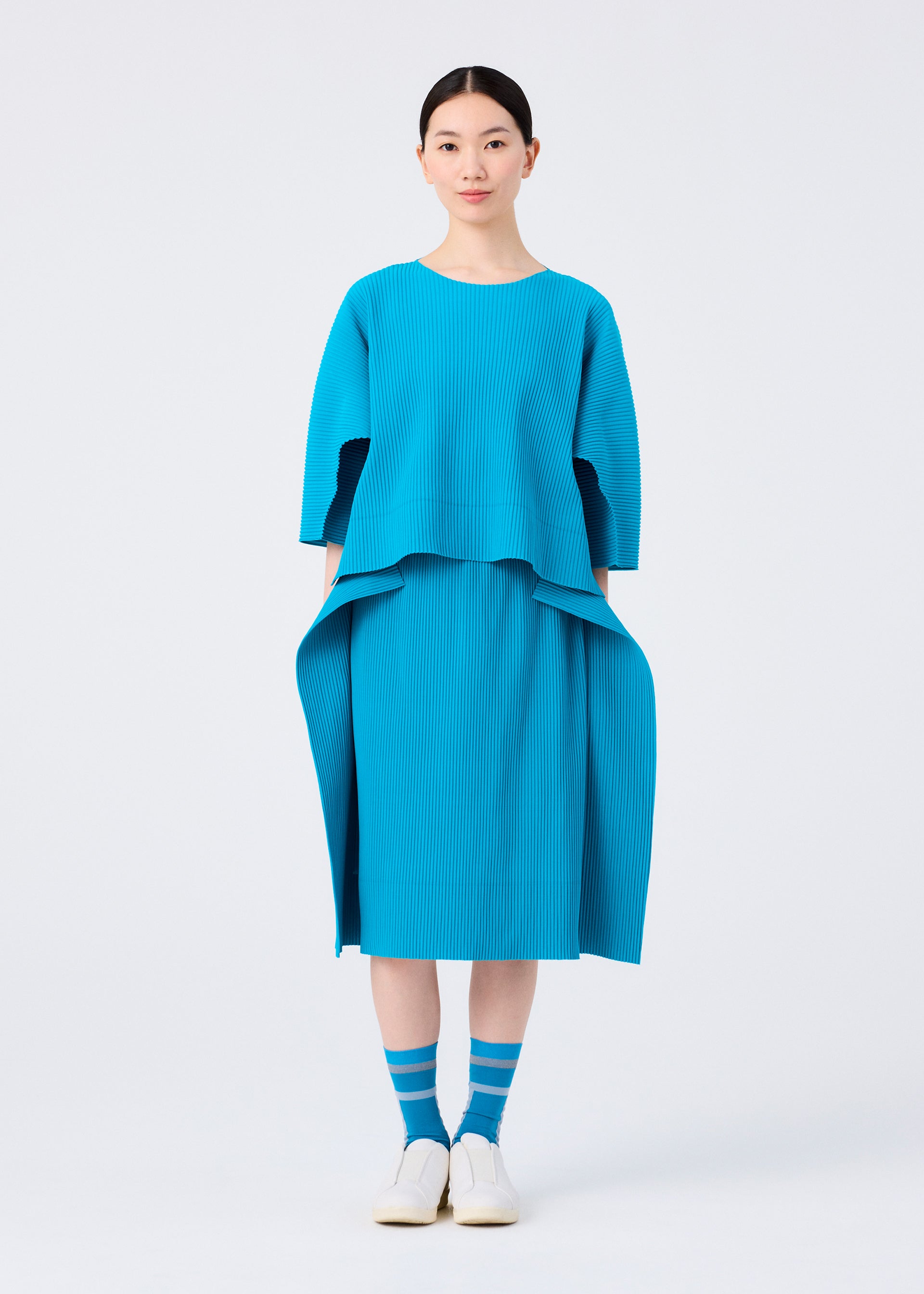 Zoomed view A woman faces forward in a me ISSEY MIYAKE TRICK PLEATS bright blue fine knit skirt with matching loose top, blue and white striped socks, and white slip-on shoes against a plain white background.