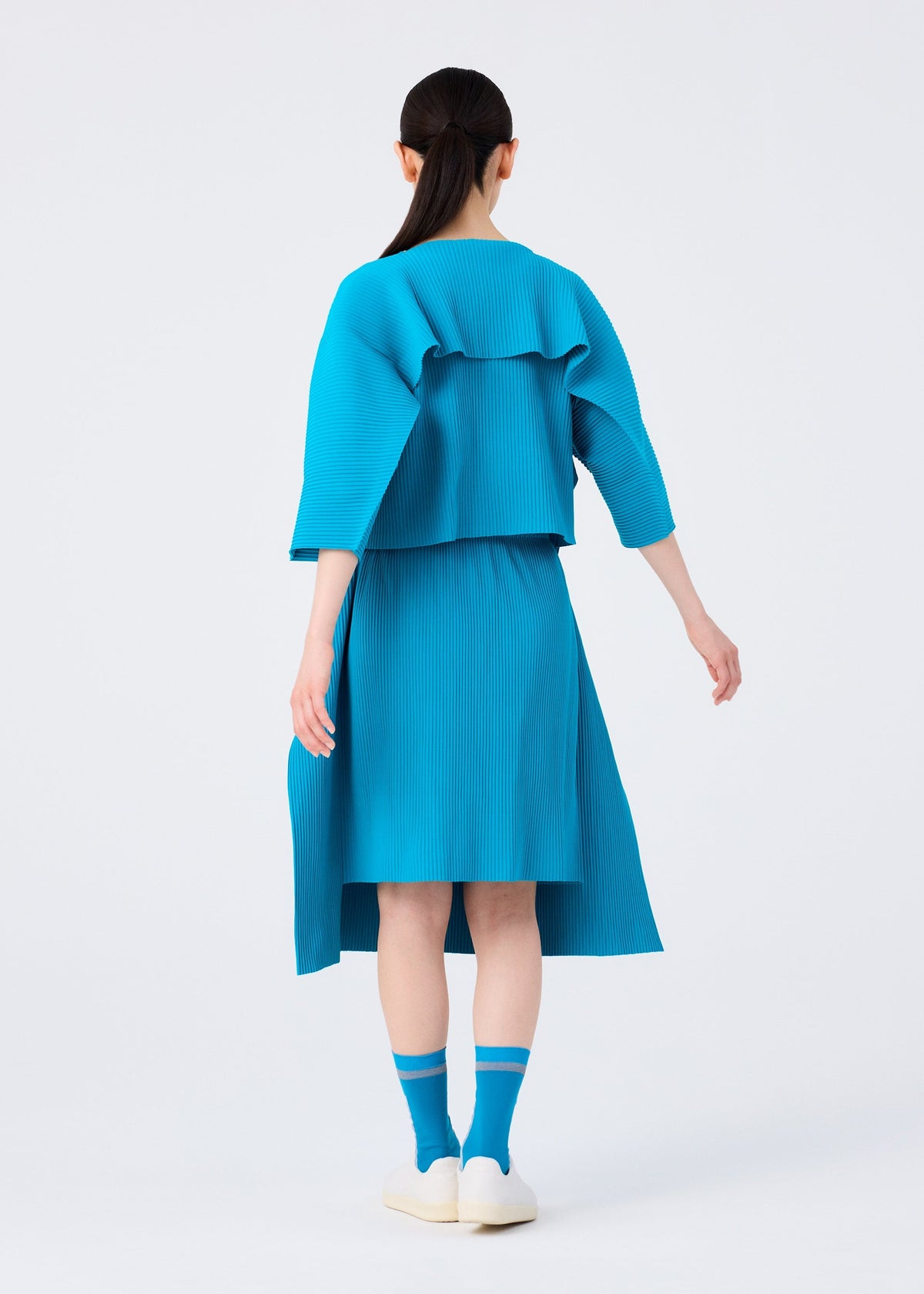 A person with long dark hair in a ponytail wears the me ISSEY MIYAKE TRICK PLEATS bright blue fine knit pleats skirt and oversized top, with matching blue socks and white shoes, standing against a plain white background, facing away. view 9 of 9