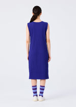 A person faces away, wearing the me ISSEY MIYAKE FINE KNIT PLEATS COLOR 1 sleeveless blue dress with purple and white striped fuzzy socks and white slip-on shoes, against a plain light background.
