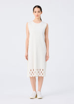 A woman faces forward wearing the APERTURE PLEATS dress by me ISSEY MIYAKE in white, featuring fine knit pleats, a textured pattern, and oval cut-outs near the hem. Her straight dark hair is pulled back and she pairs it with white slip-on shoes.