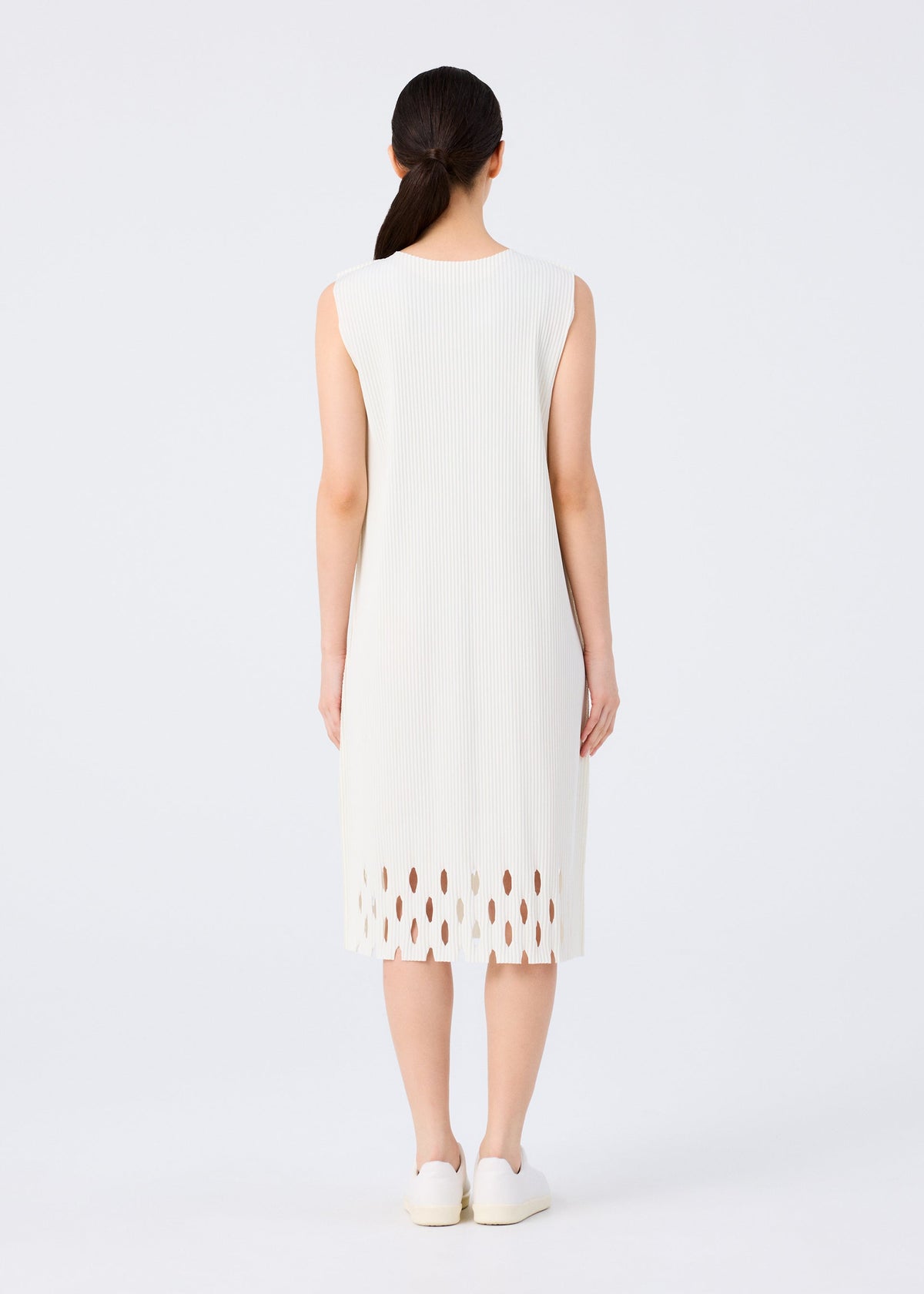 A woman with long dark hair in a low ponytail, facing away, wears the me ISSEY MIYAKE APERTURE PLEATS sleeveless knee-length pleated dress with a cutout hem and white shoes, standing against a plain white background. view 6 of 6