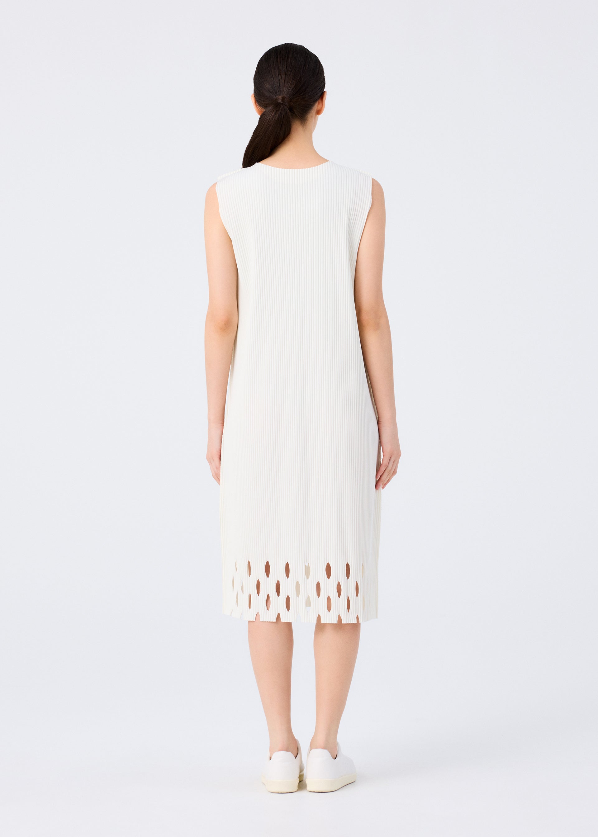 Zoomed view A woman with long dark hair in a low ponytail, facing away, wears the me ISSEY MIYAKE APERTURE PLEATS sleeveless knee-length pleated dress with a cutout hem and white shoes, standing against a plain white background.