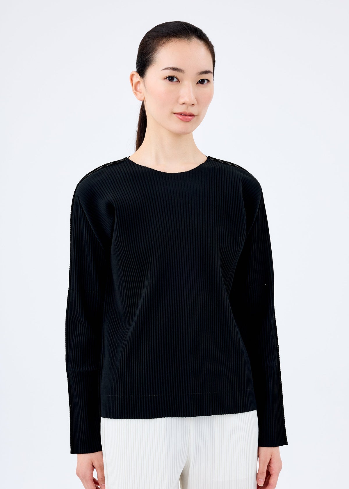 A woman with long dark hair tied back is wearing the FINE KNIT PLEATS BLACK top by me ISSEY MIYAKE with light-colored pants, standing against a plain white background facing forward with a neutral expression. view 3 of 5