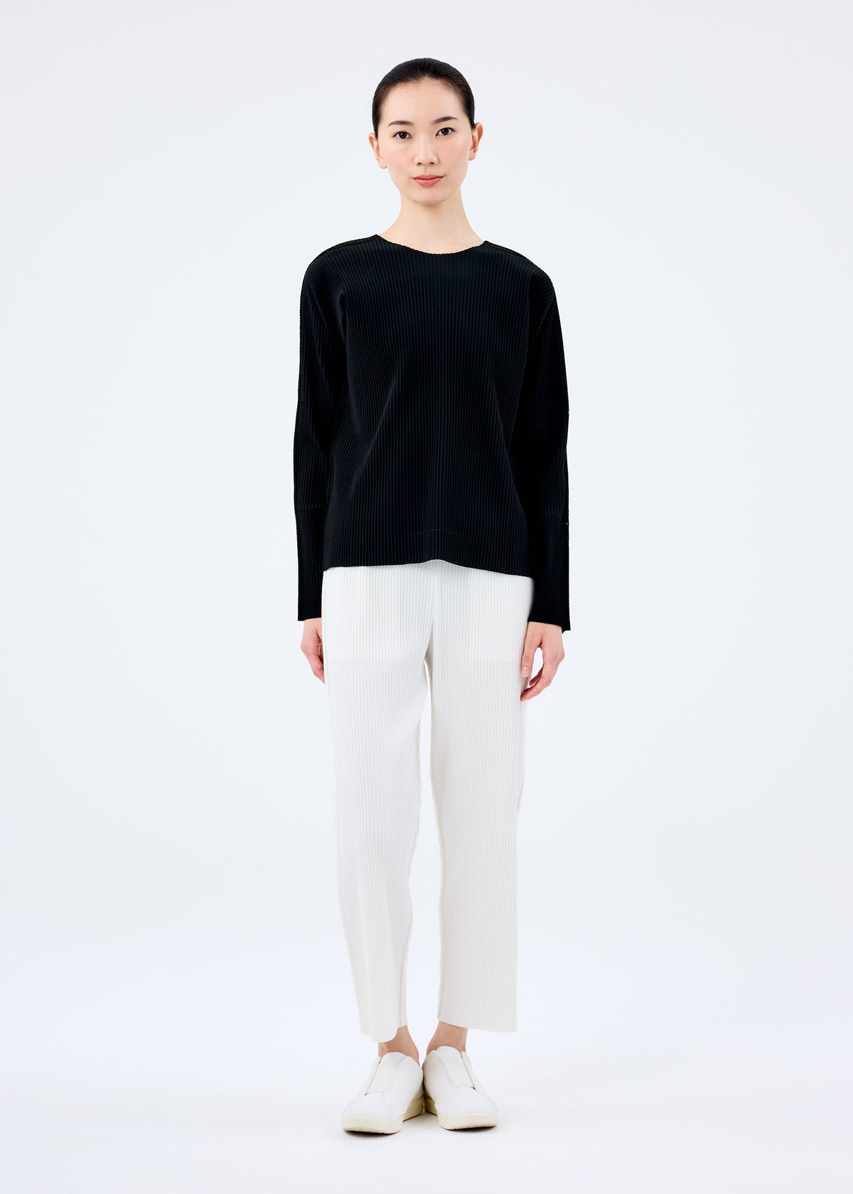 A woman faces forward against a white background, wearing the "FINE KNIT PLEATS BLACK" top by me ISSEY MIYAKE, paired with white cropped pants and white slip-on shoes. view 5 of 5