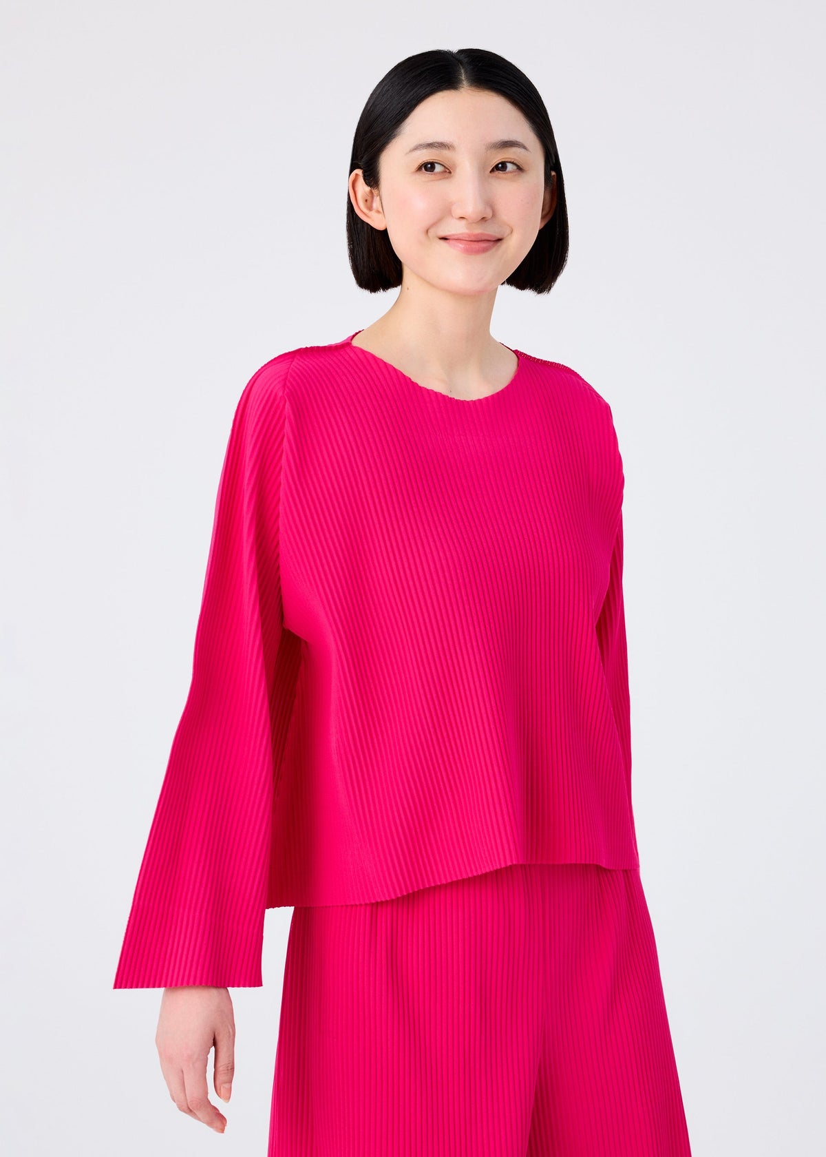 A woman with short black hair is smiling, wearing the FLARE PLEATS set by me ISSEY MIYAKE—a bright pink, long-sleeved, finely pleated top and matching pants—standing against a plain light background. view 6 of 8