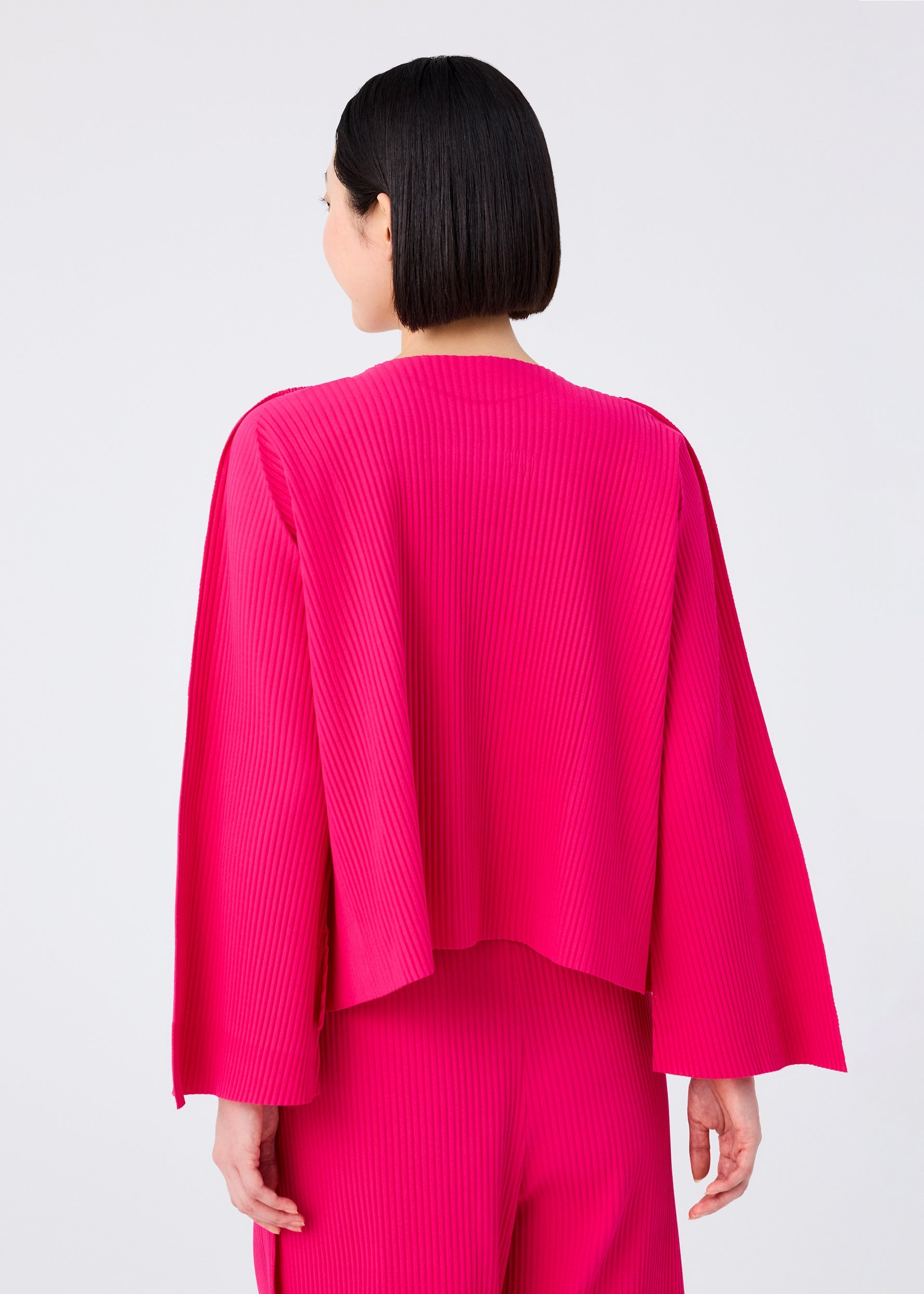 Zoomed view A person with short dark hair, seen from behind, wears the FLARE PLEATS set by me ISSEY MIYAKE—a bright pink, fine knit, long-sleeved top and matching flared pants—against a plain light background.