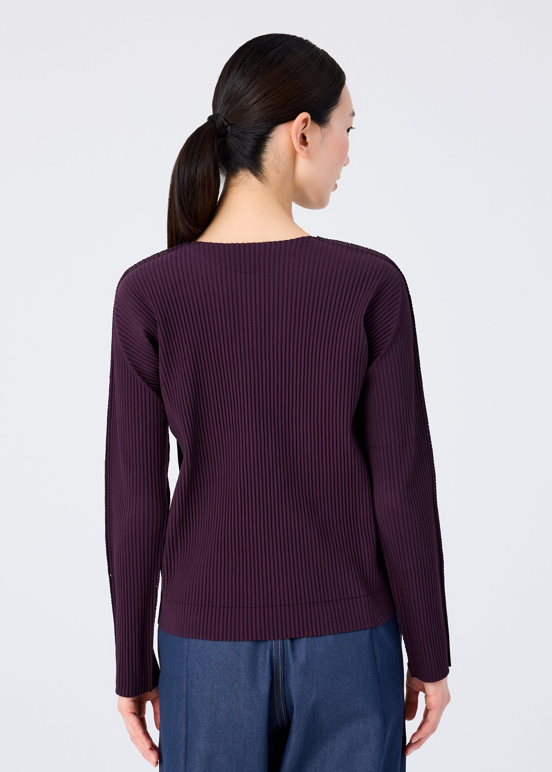 Zoomed view A woman with straight dark hair in a low ponytail is seen from behind, wearing the me ISSEY MIYAKE FINE KNIT PLEATS COLOR 1 round-neck, long-sleeved top and dark blue jeans against a plain light background.