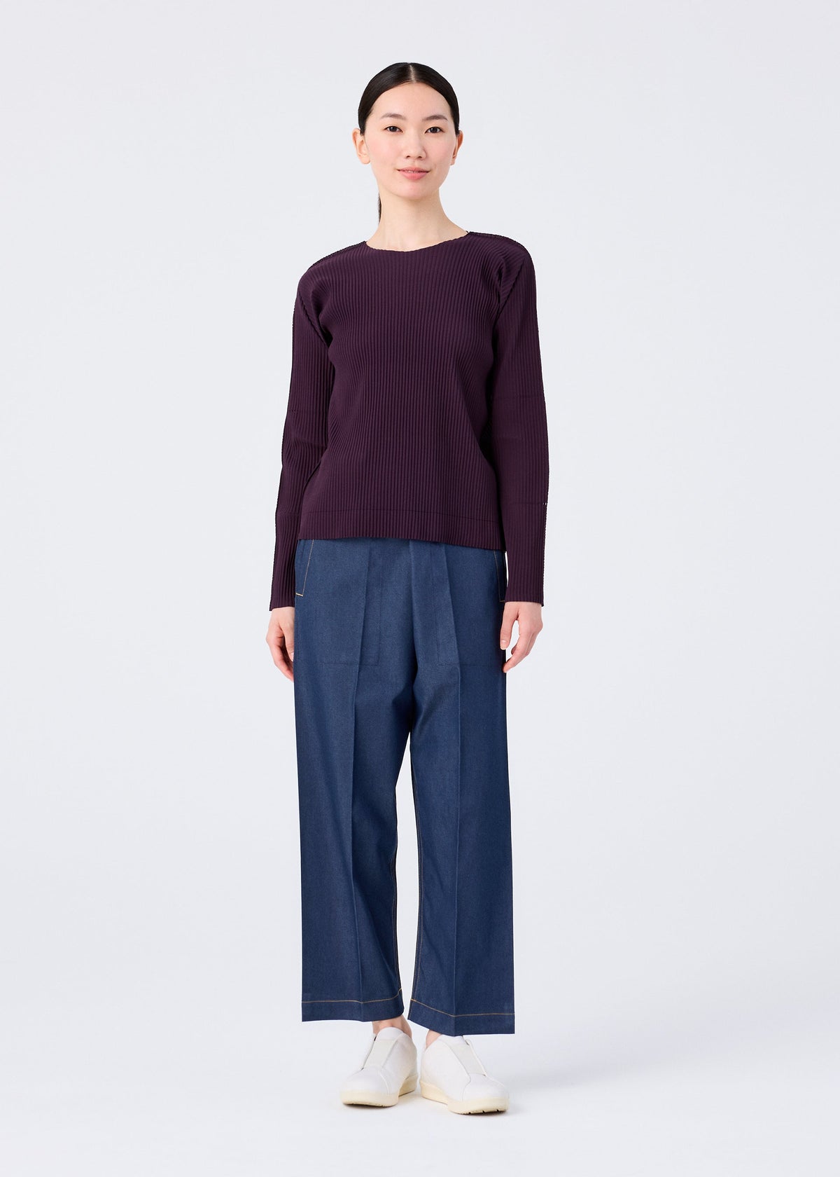 A woman faces forward against a white background, wearing the me ISSEY MIYAKE FINE KNIT PLEATS COLOR 1 long-sleeved dark purple top, wide-leg blue pants, and white slip-on shoes. view 8 of 8