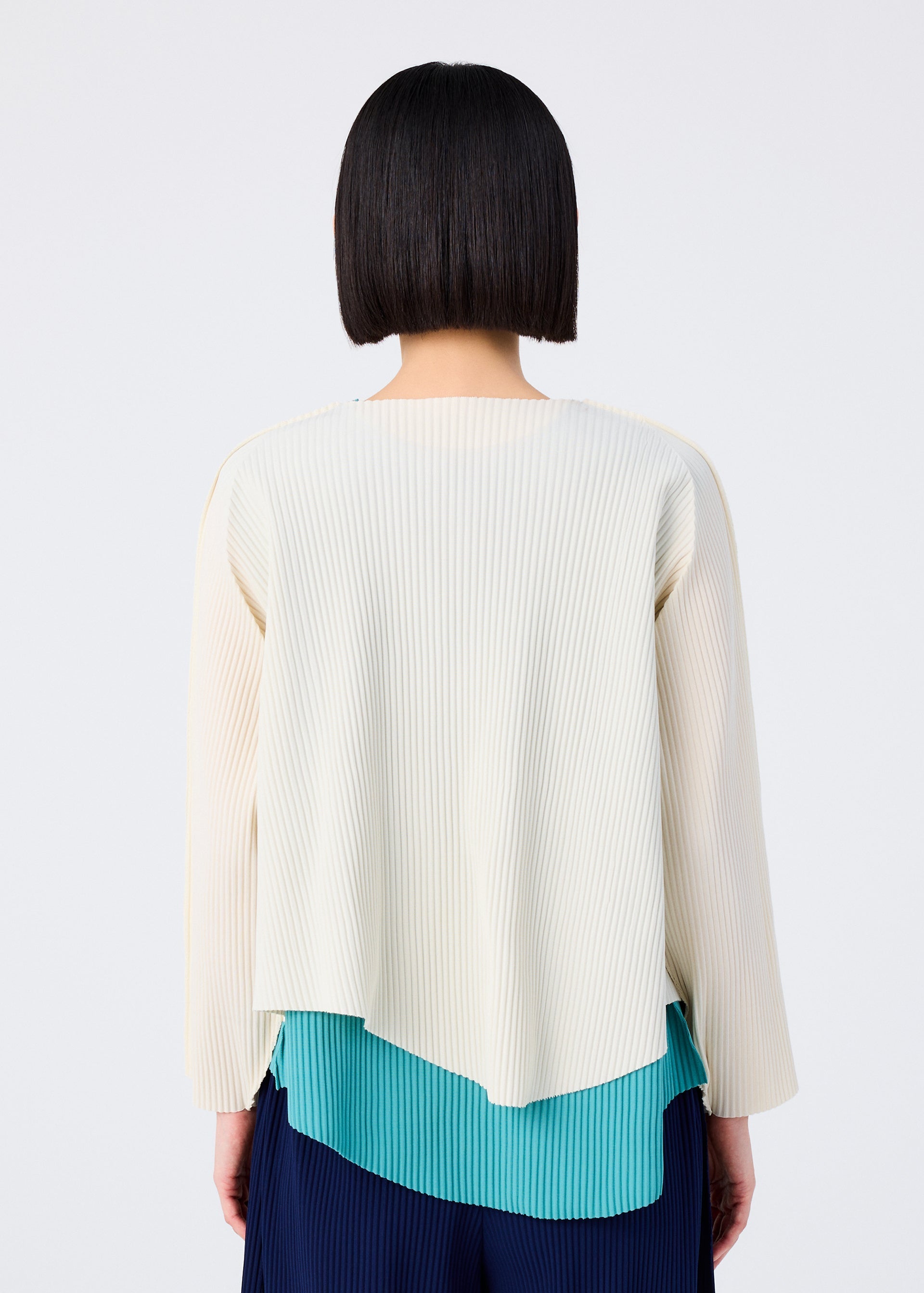Zoomed view A person with straight, chin-length dark hair is seen from behind, modeling the AURORA PLEATS set by me ISSEY MIYAKE—a lightweight, finely pleated long-sleeved top layered over a teal pleated shirt and navy pleated pants.