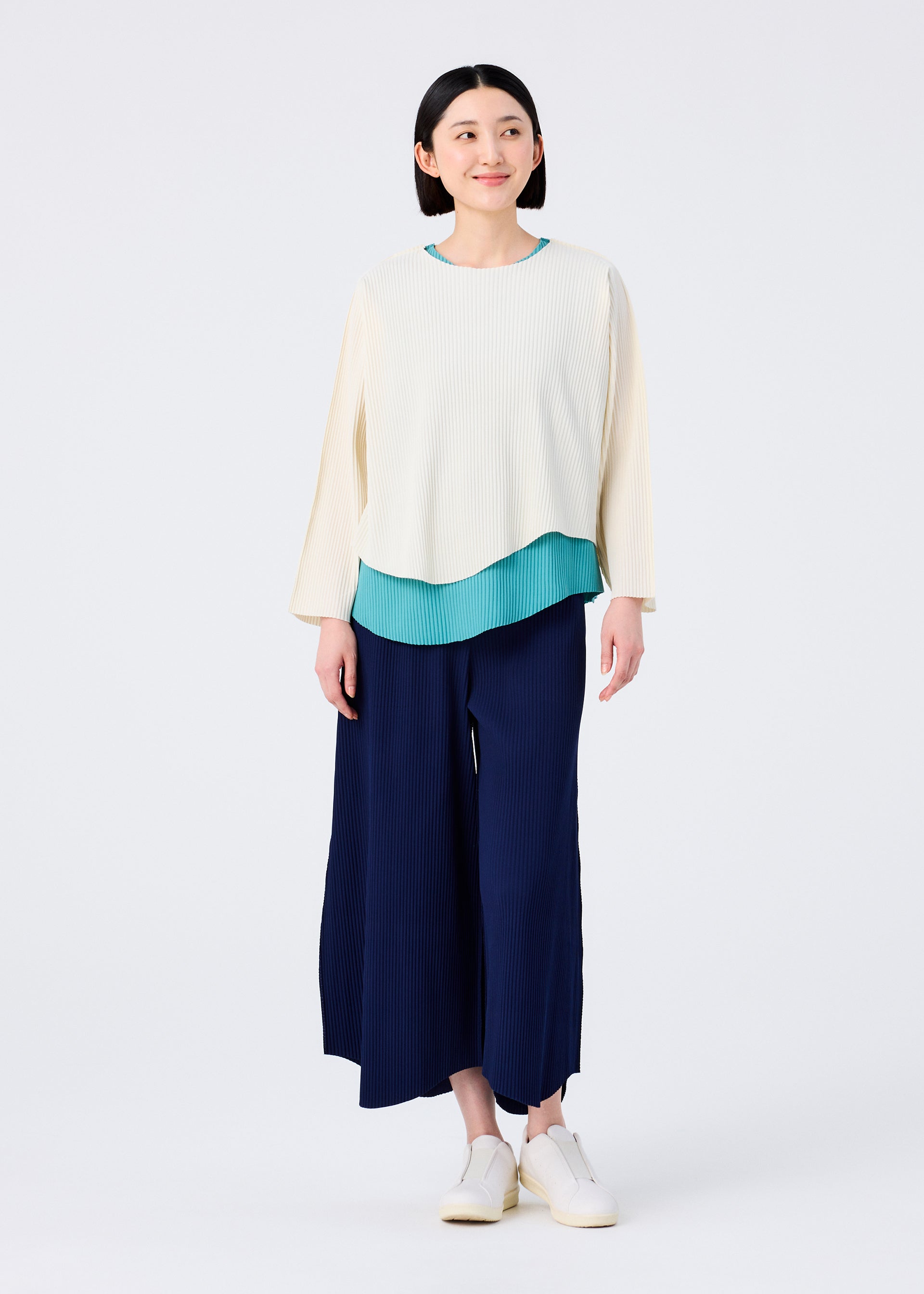 Zoomed view A woman with straight dark hair and a slight smile wears the AURORA PLEATS wide navy pants by me ISSEY MIYAKE, paired with a lightweight long-sleeved top over a teal blouse and white slip-on shoes, standing against a plain background.