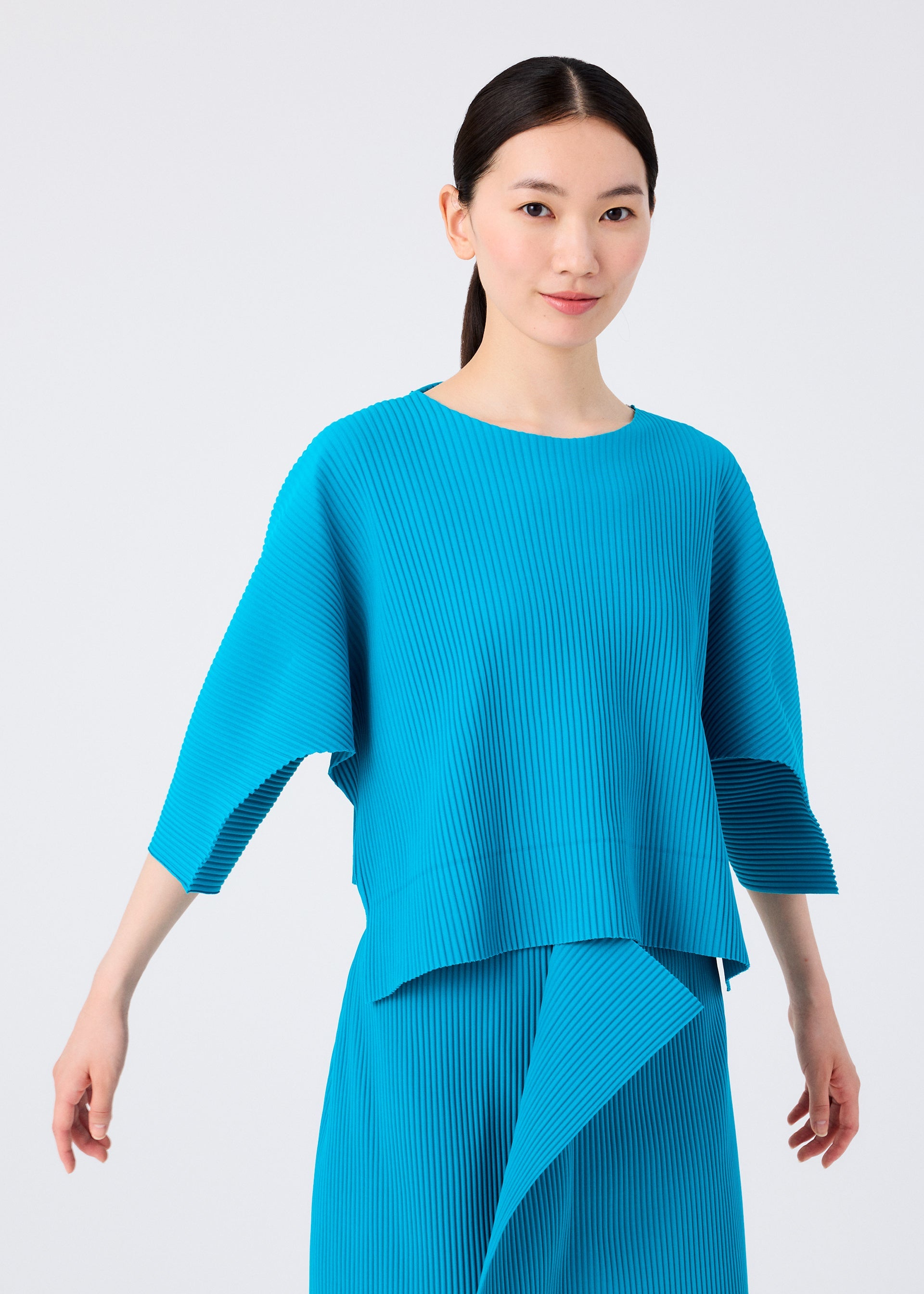 Zoomed view A woman with straight black hair models the TRICK PLEATS set by me ISSEY MIYAKE—a bright blue, loose-fitting, fine knit pleated top and matching skirt—standing gracefully with arms outstretched against a white background.